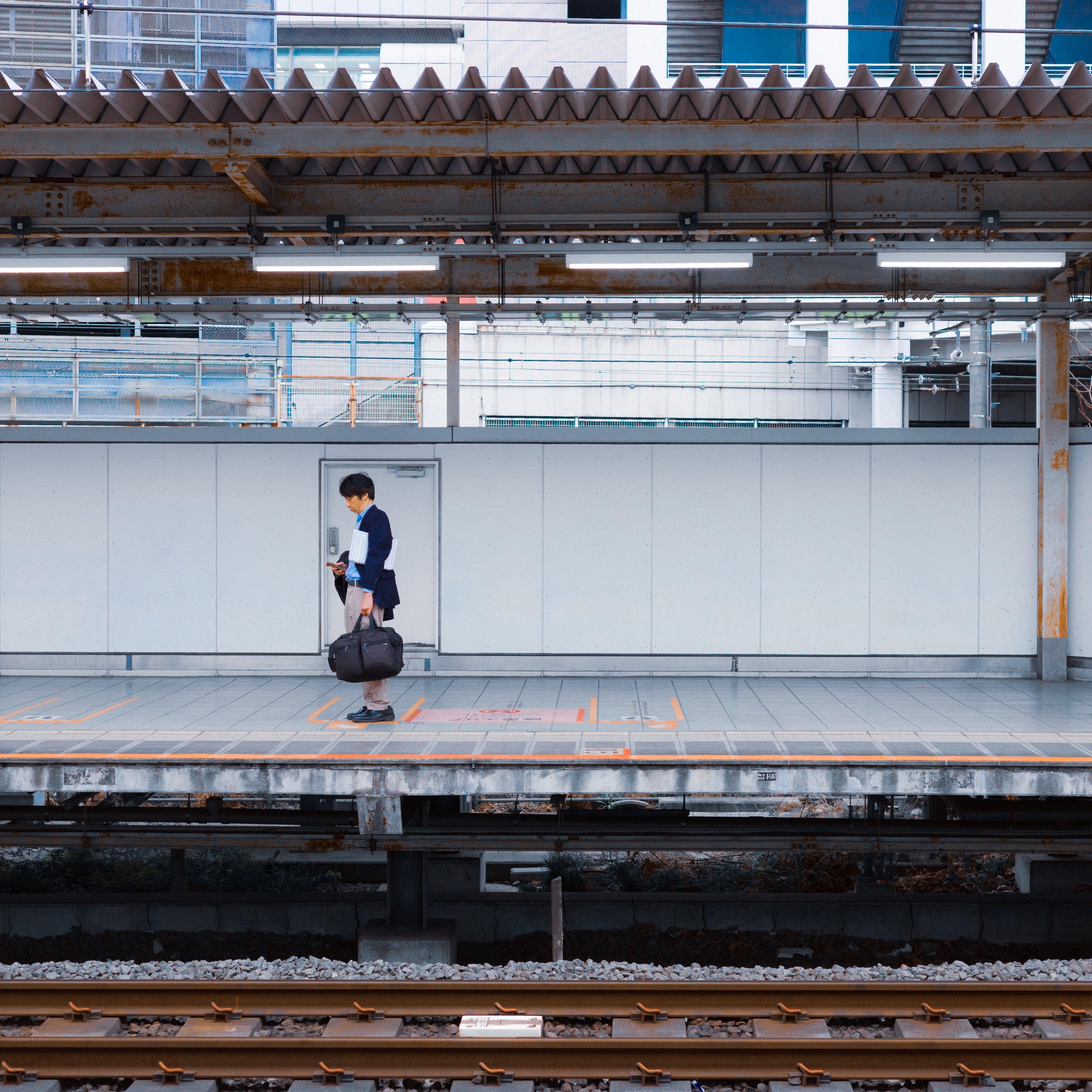 The image shows a person standing on a railway platform holding a bag. The platform has a modern, industrial look with visible overhead structures. There are train tracks in the foreground, and the background features urban architecture.