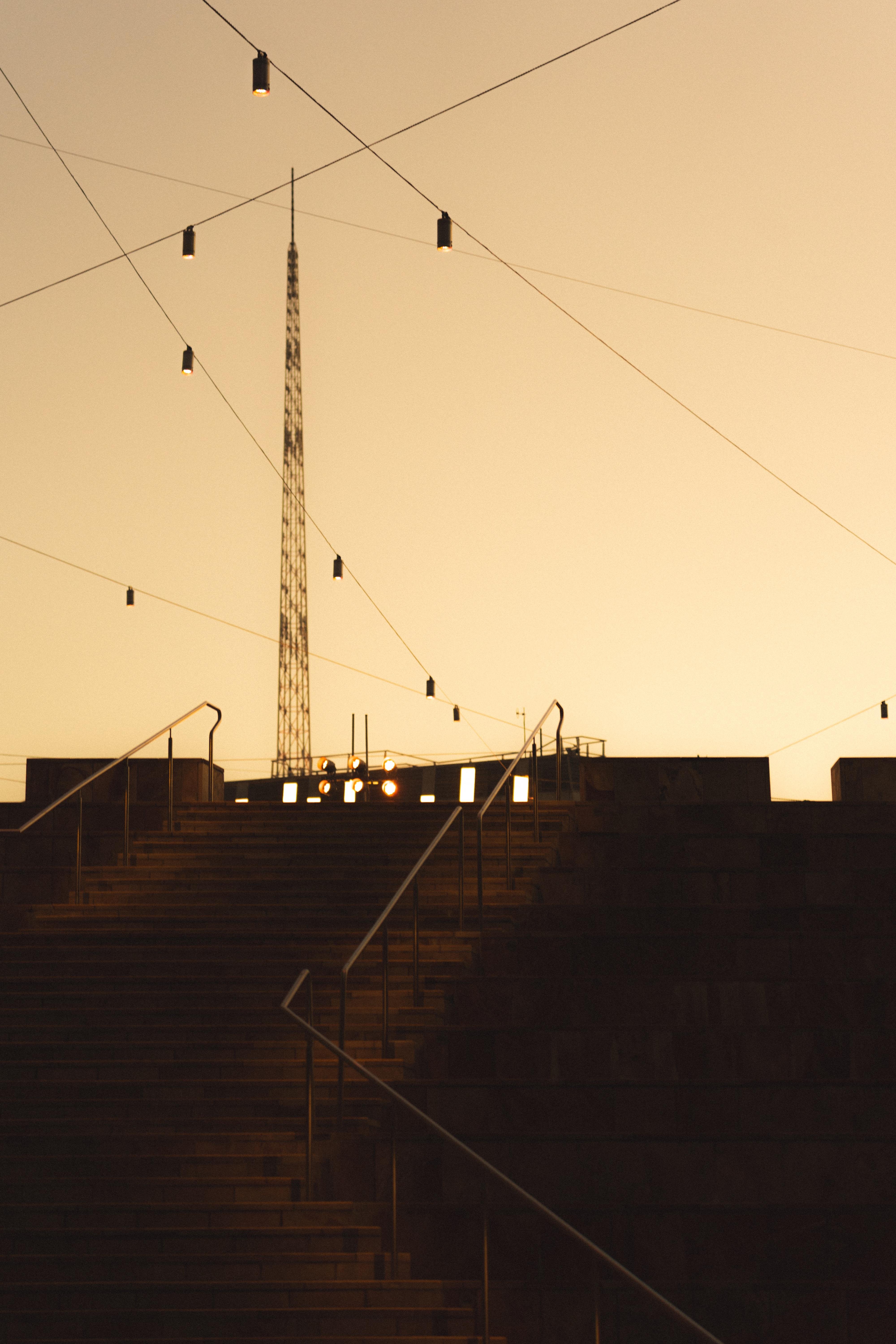 Wide stone staircase leading up to a platform with railings. Overhead, several strings of hanging lights crisscross against the sky. A tall, slender communication tower is visible in the background, silhouetted by a warm, golden sunset.