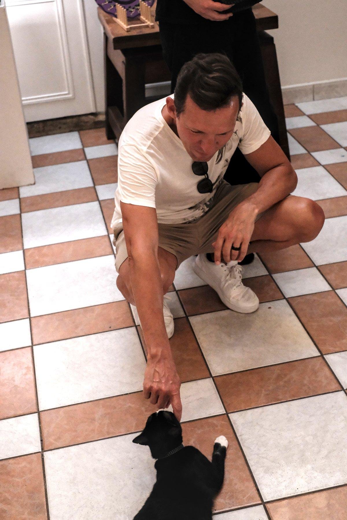 Photo verticale. Sur un sol en carrelage blanc et marron, un homme est accroupi et tends sa main droite vers un chat noir couché au sol qui renifle la main tendue