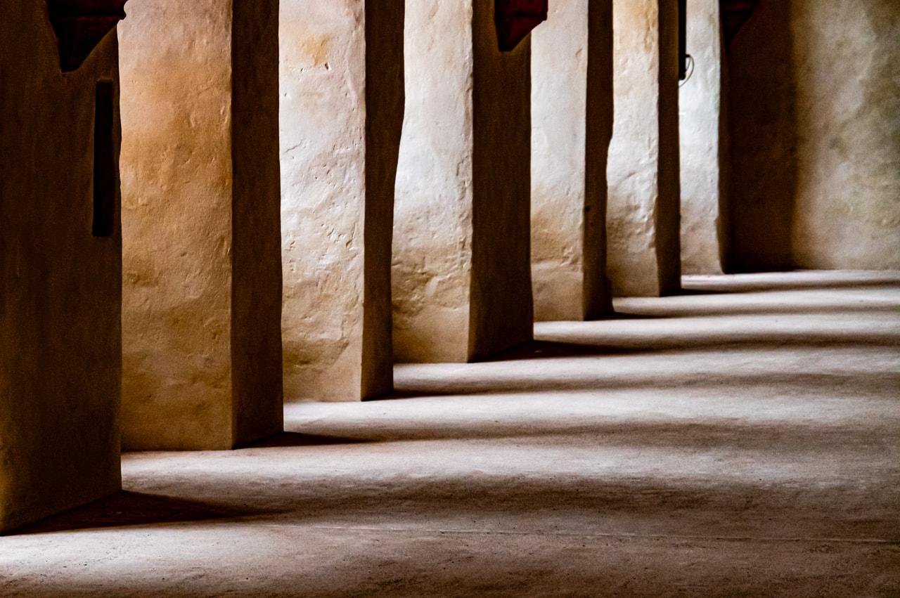 This image shows a row of architectural columns or pillars casting shadows on the ground. The lighting creates a pattern of light and shadow, highlighting the texture of the walls. It seems to be an interior space with a rustic or historical feel.