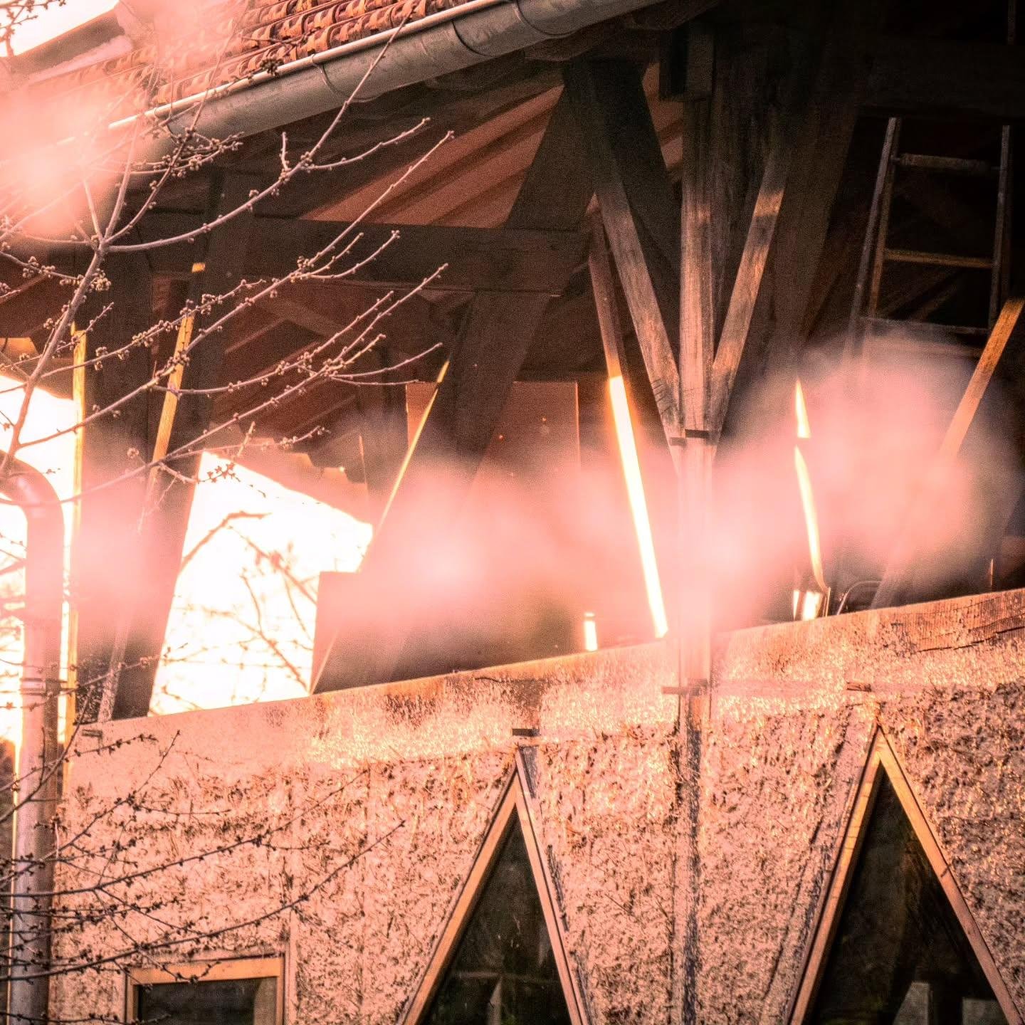 The image shows part of a building with wooden beams and a roof, possibly a rustic or cabin-like structure. There are some bare tree branches in the foreground and a textured wall with triangular elements in the design. The lighting gives the scene a warm, glowing effect, suggesting sunset or sunrise.