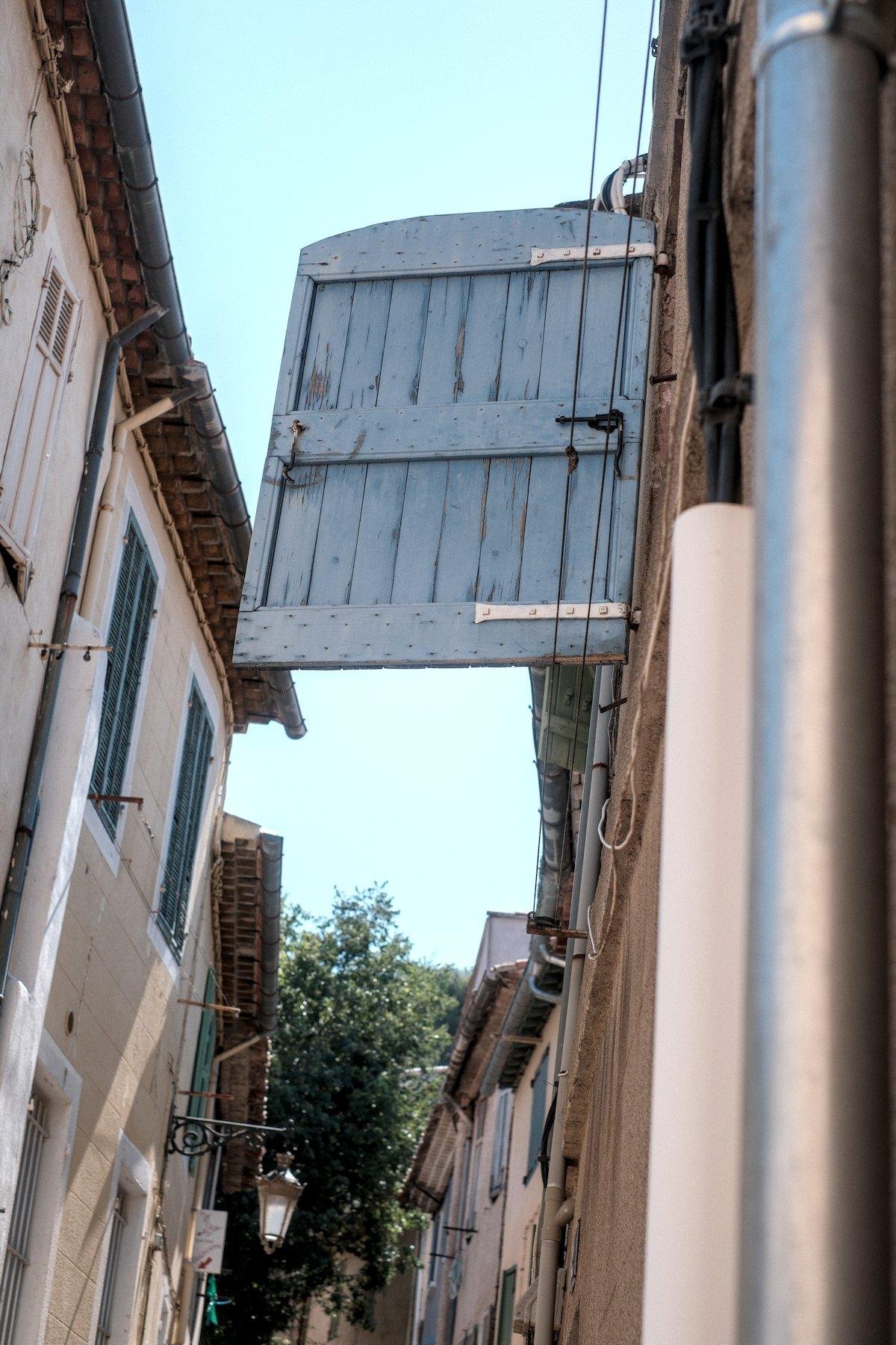 Photo verticale. Dans une rue étroite, un grand volet est perpendiculaire à son mur et semble dessiner une porte en plein vide
