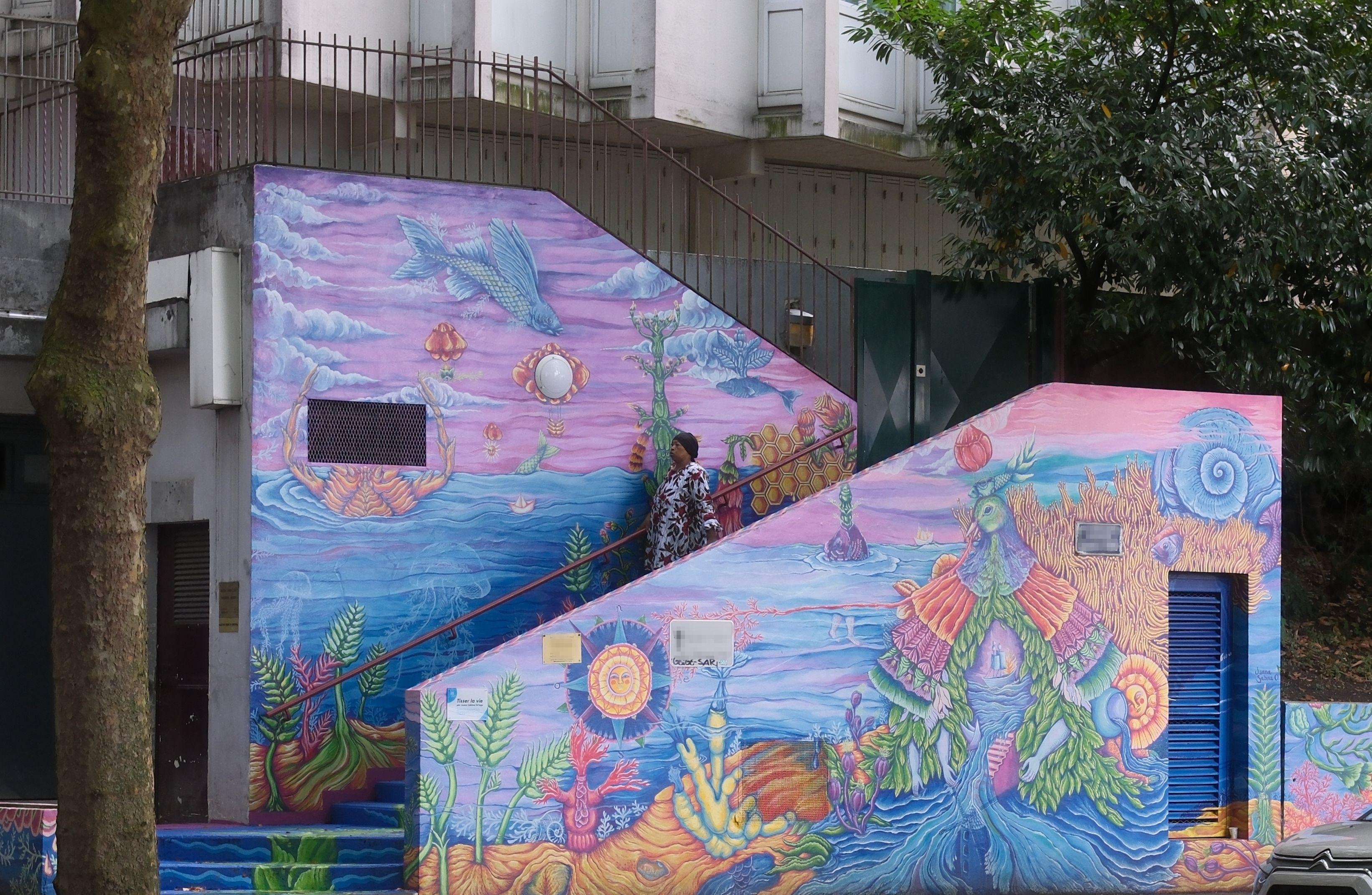 A photo of an outside stairway going right then change to left mid way. The walls of the stairway are heavily painted with multiple colours. A woman is going down