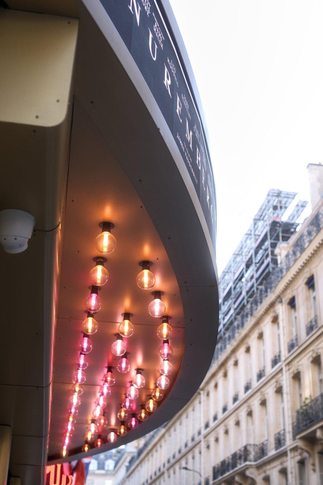 Photo verticale. Sur la gauche, des ampoules de style ancien de diverses teintes rouges occupant le dessous d’un petit toit de devanture de cinéma. Sur la droite, un immeuble parisien typique intra muros quartier chic