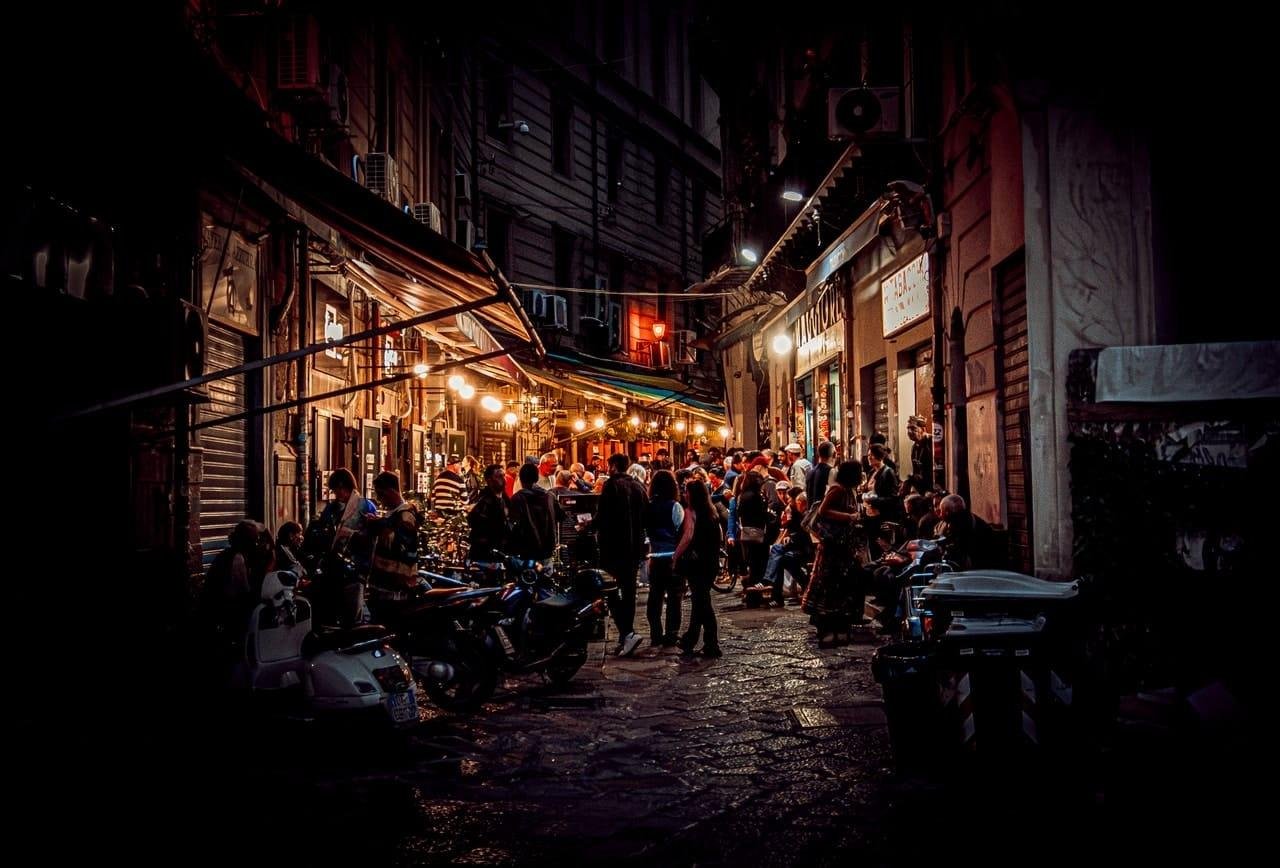 Crowded narrow street at night with people gathered outside small bars and shops under striped awnings and warm hanging lights; scooters and motorcycles parked along the cobblestone lane between tall, closely spaced buildings.