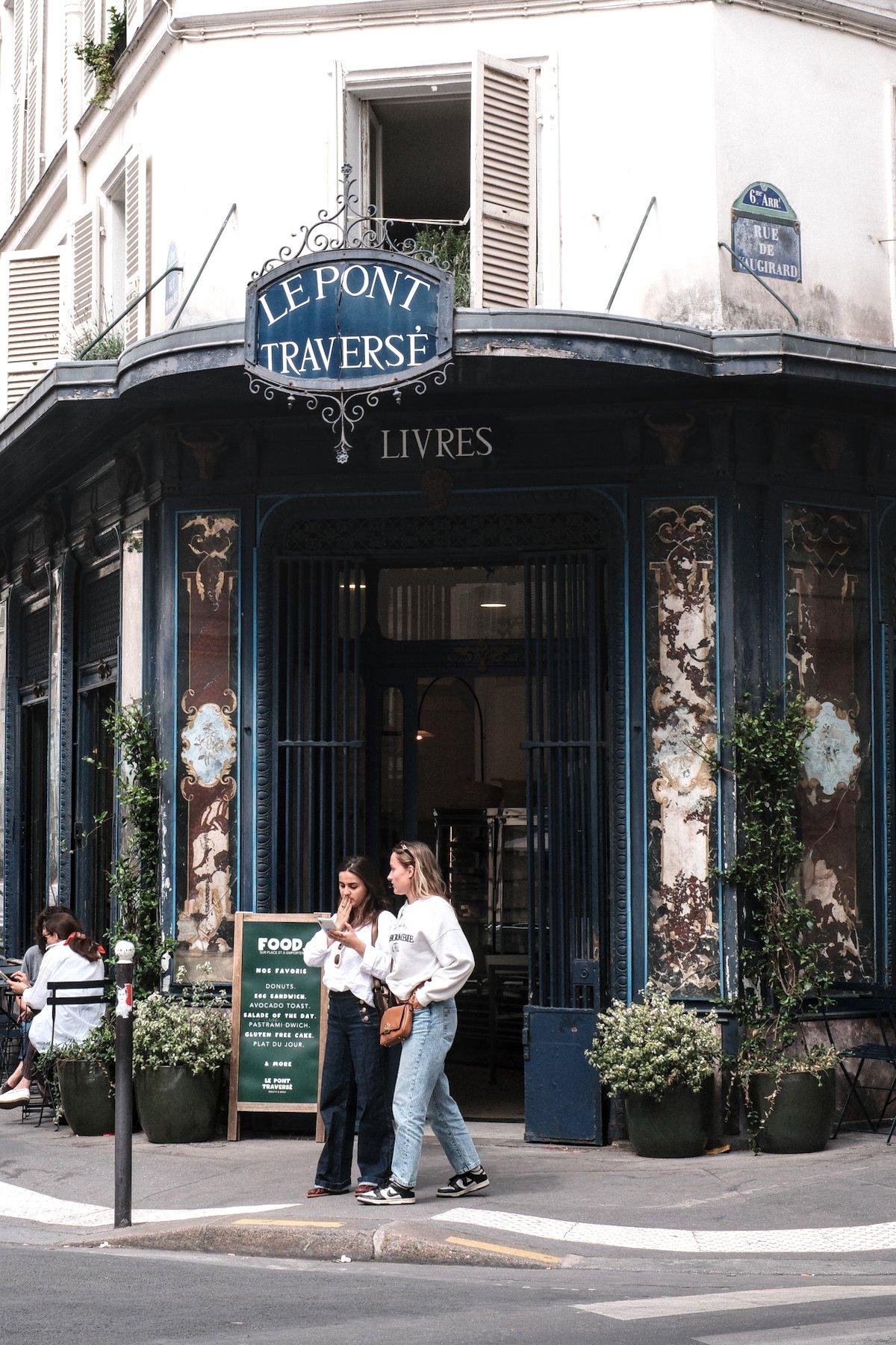 Photo verticale. À l’angle d’une rue, l’entrée bleue de l’établissement « Le pont traversé » écrit sur une enseigne aux bords en fer forgé, accroché à un petit toit. Dessous, au dessus de la porte d’entrée on peut lire « Livre ». Devant l’entrée, une ardoise sur pied annonce le menu du brunch. Sur la droite, 2 jeunes femmes passent