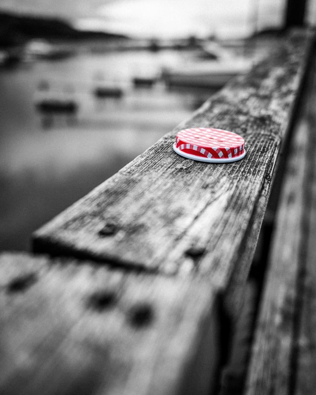 The image shows a close-up of a red and white checkered jar lid on a wooden surface. The lid is in color while the rest of the image is in black and white. The background appears to be a blurred waterfront scene with boats.