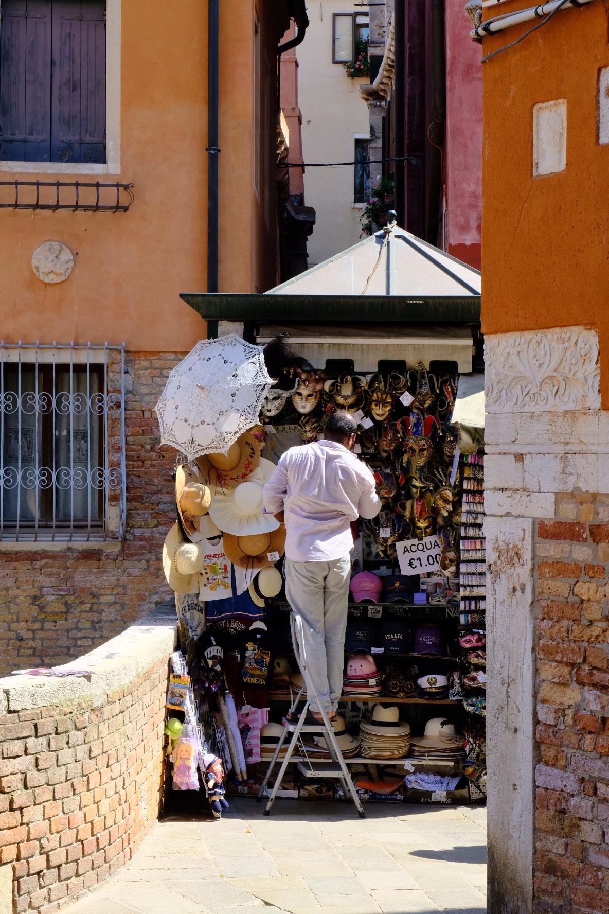 A merchant standing on a small step ladder at an outdoor souvenir kiosk in a narrow alley, with shelves of Venetian-style masks and other souvenirs; white lace parasol and straw hats hang on the left, and a sign reads “ACQUA €1.00,” framed by orange and red building walls and a brick walkway.