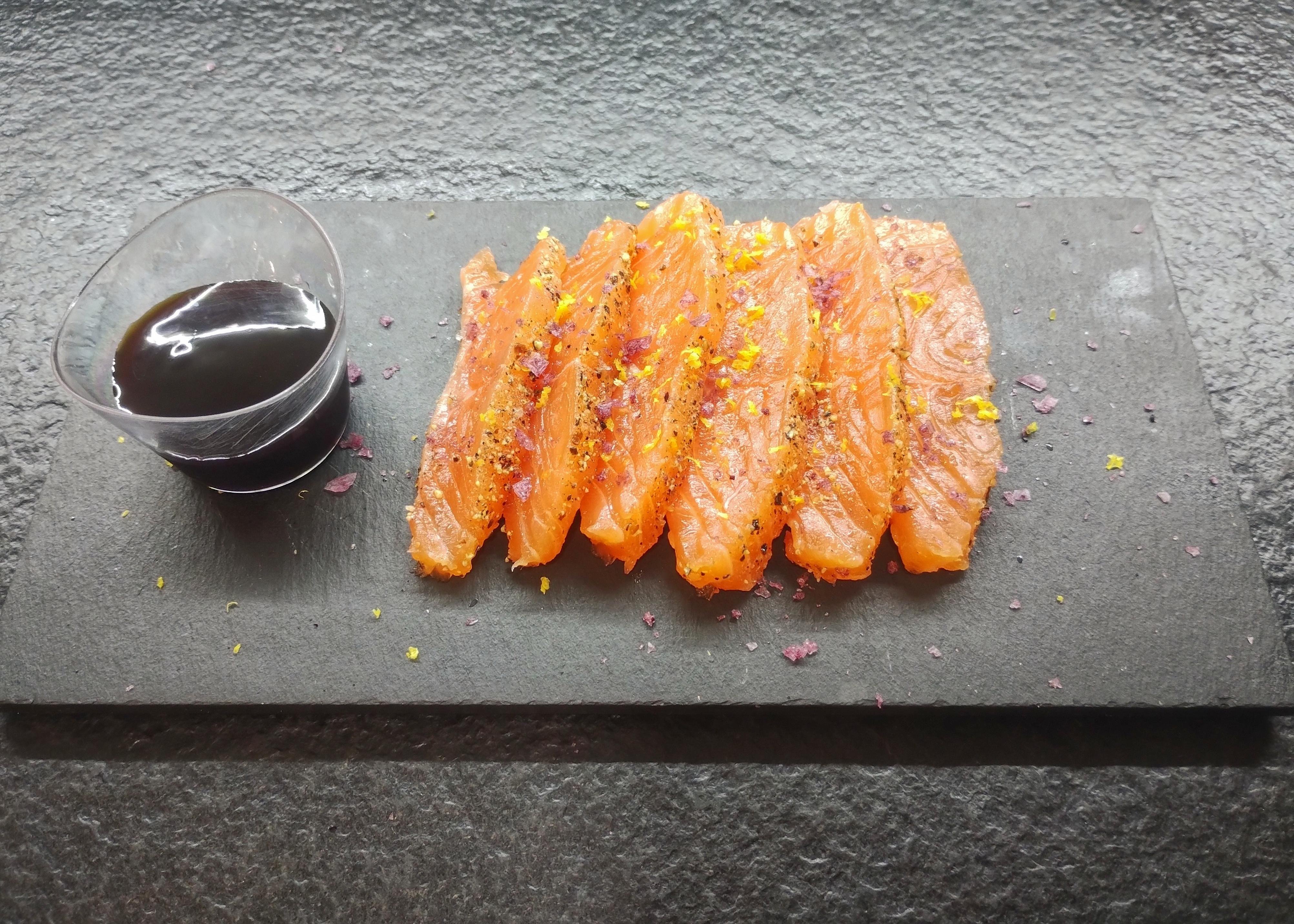 The image shows slices of raw salmon on a dark serving platter. They are garnished with what appears to be seasoning such as pepper and lemon zest. There is also a small container of dark dipping sauce, likely soy sauce, next to the salmon.