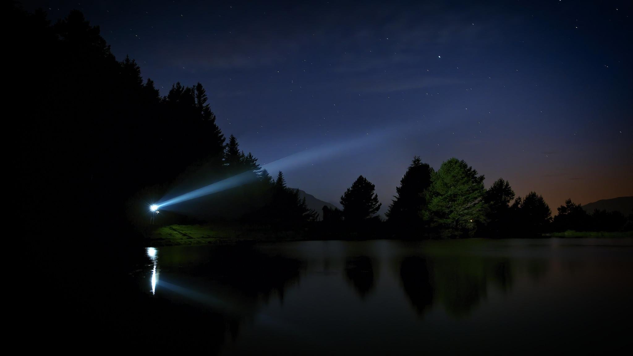 This image shows a nighttime scene with a body of water reflecting trees and the night sky. A beam of light, likely from a flashlight or torch, is shining from the left side, illuminating part of the landscape. The sky is clear, showcasing stars scattered across it. The overall atmosphere is calm and serene.