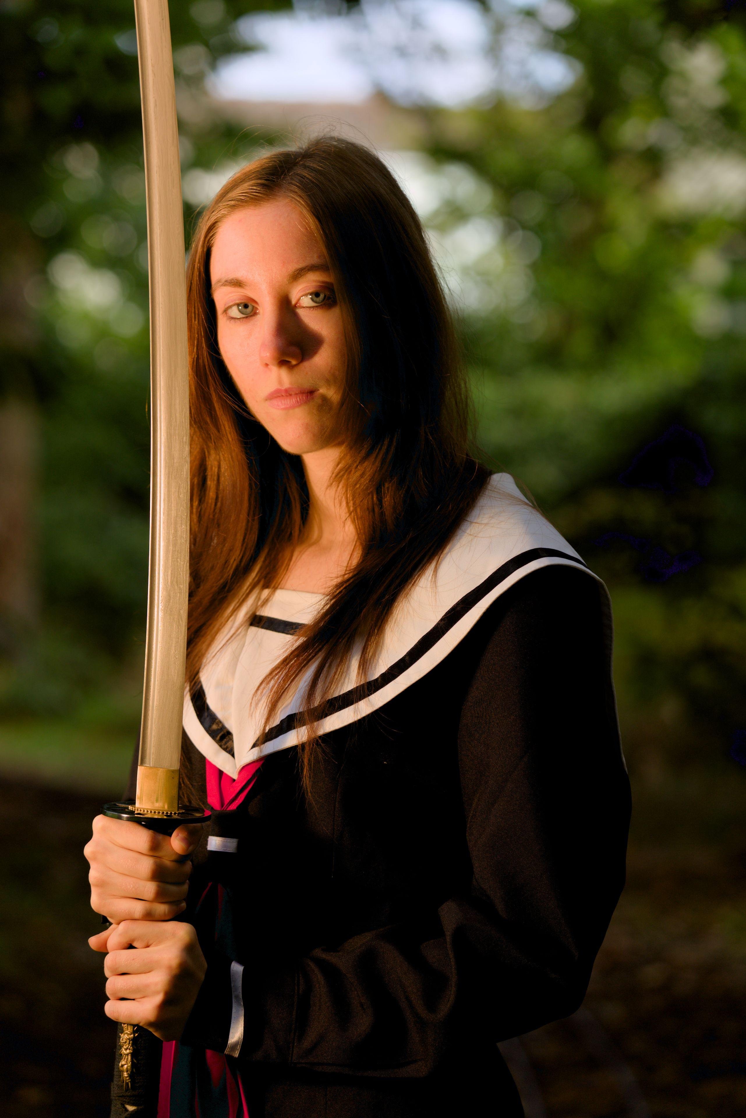 A young red hair woman wearing a Japanese style school uniform is holding a katana vertically near her head, looking at the camera with a menacing glaze. An orange light is coming from the left and the background shows some blurry vegetation
