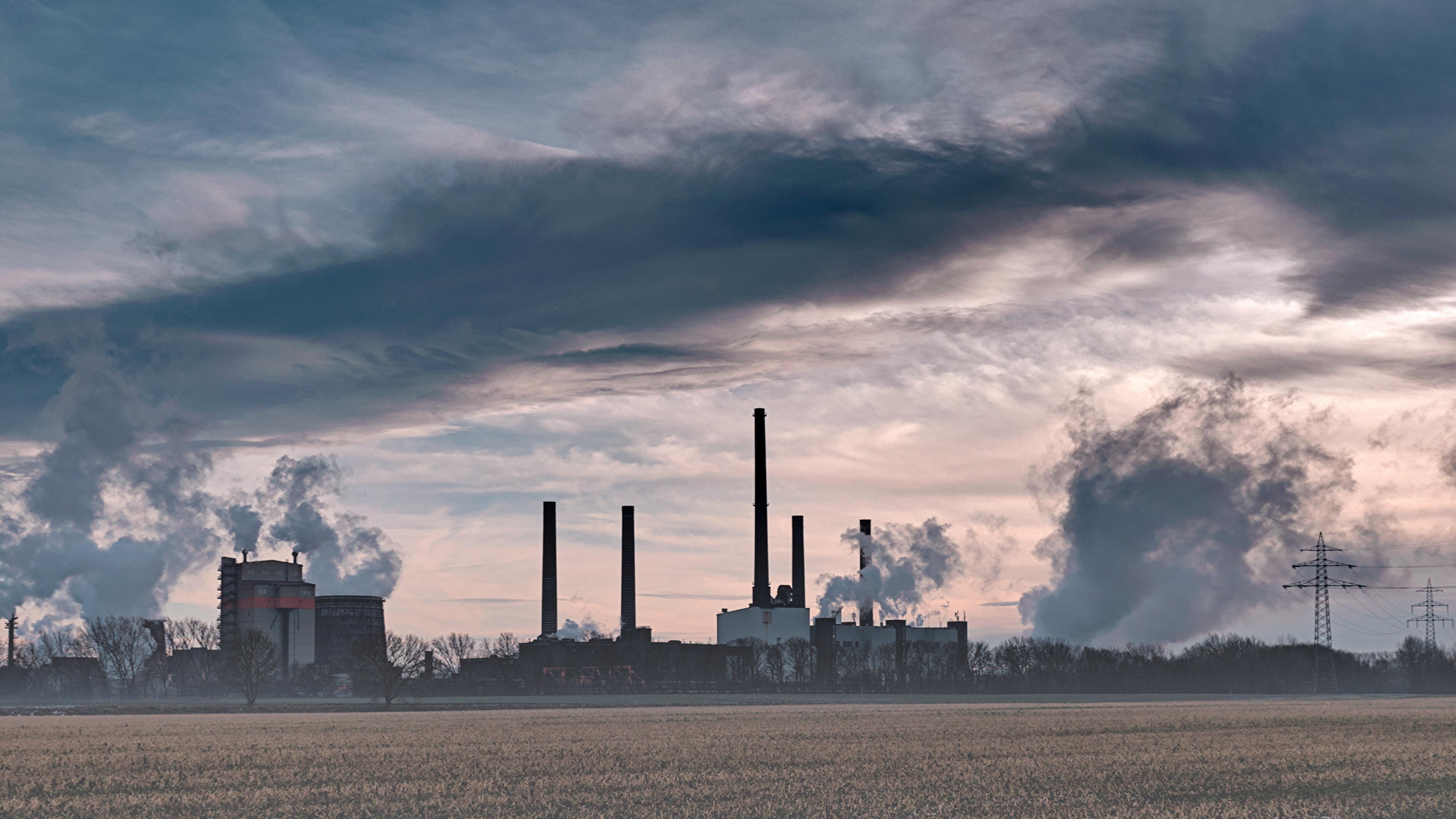 The image depicts an industrial scene with several factories or power plants characterized by tall smokestacks emitting smoke or steam into the sky. The sky is cloudy, and in the foreground, there is an open field, suggesting the location might be on the outskirts of a city or in an industrial area. Power lines are visible as well, indicating the presence of electrical infrastructure.