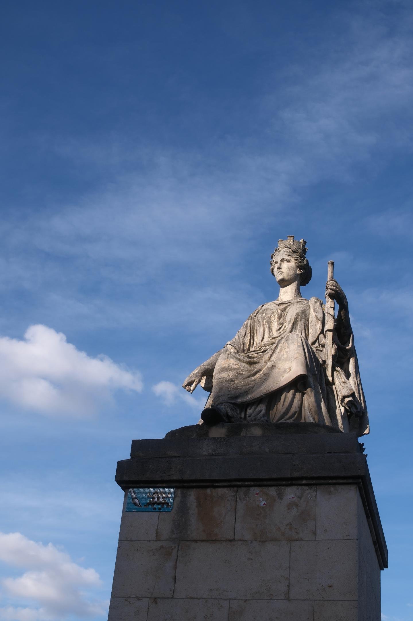 Photo verticale. Une statue au sommet d’un piédestal. Elle prends le soleil alors que le socle est dans l’ombre, sur fond de ciel bleu tâché de nuages