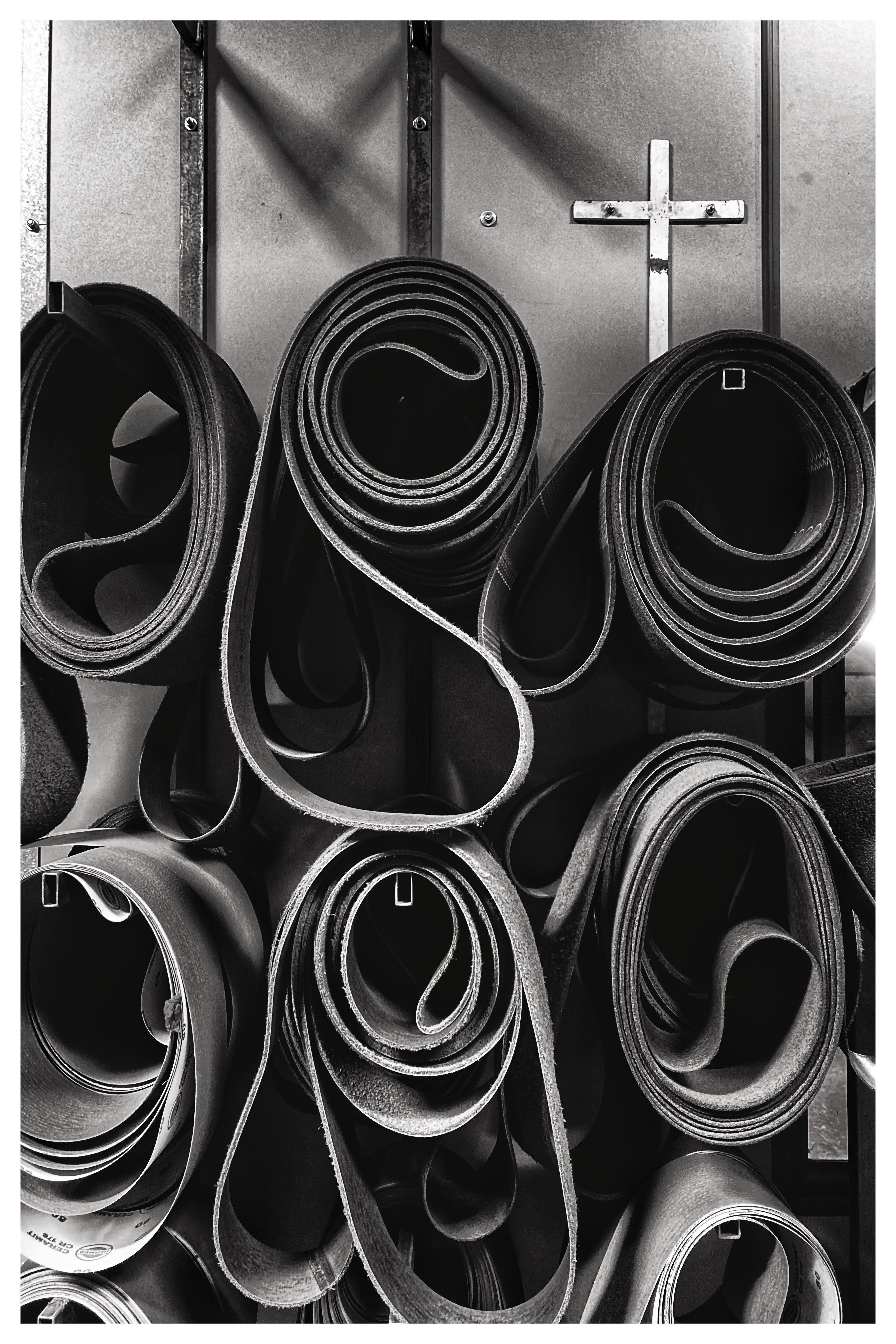 Black-and-white close-up of multiple rolled belts or straps stacked upright against a metal wall, with a small cross mounted near the upper right.
