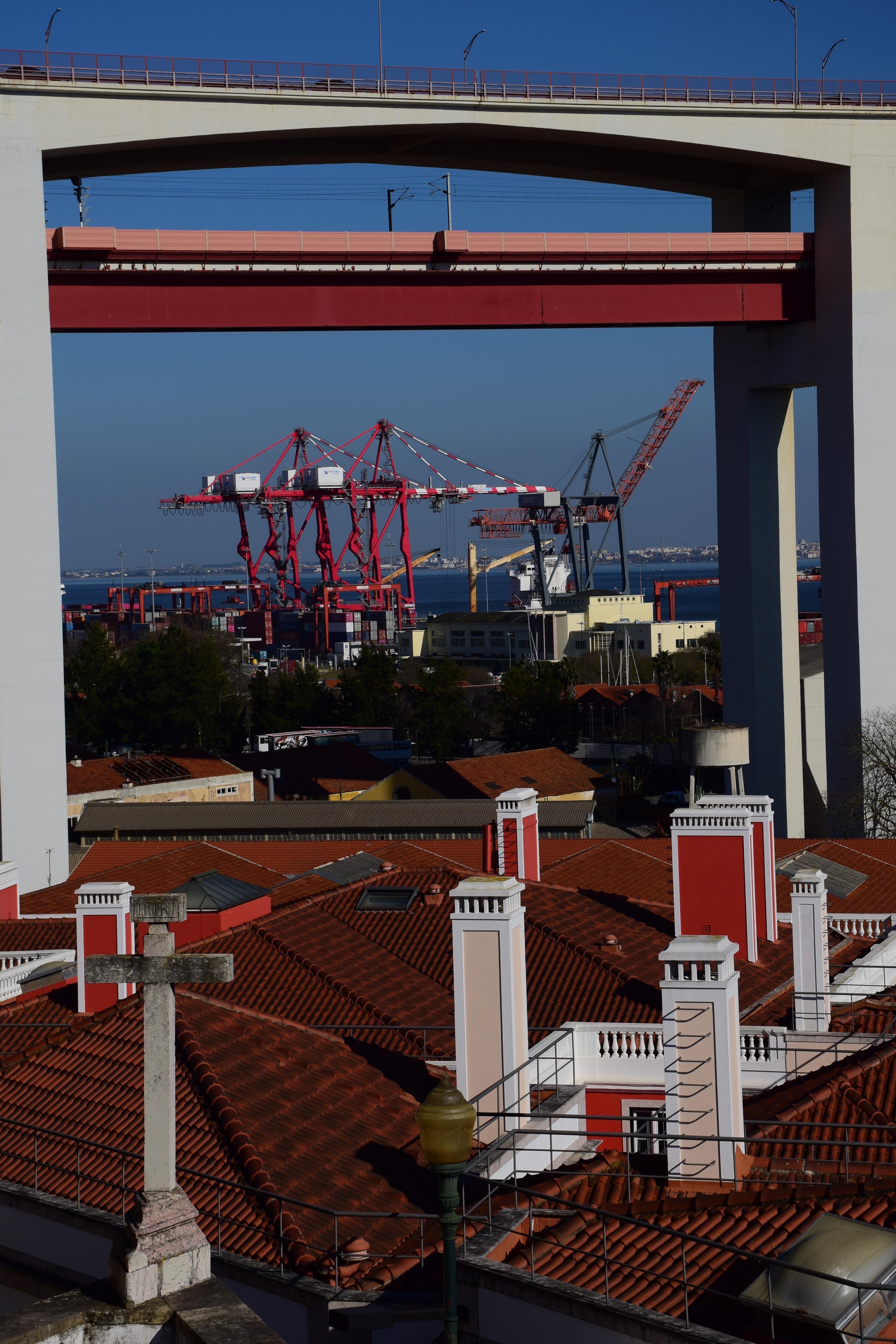 The image shows a scene with a bridge in the upper part and a view of a harbor with cranes in the background. In the foreground, there are buildings with red tile roofs and white chimneys. The setting suggests an urban area near a port or industrial zone.