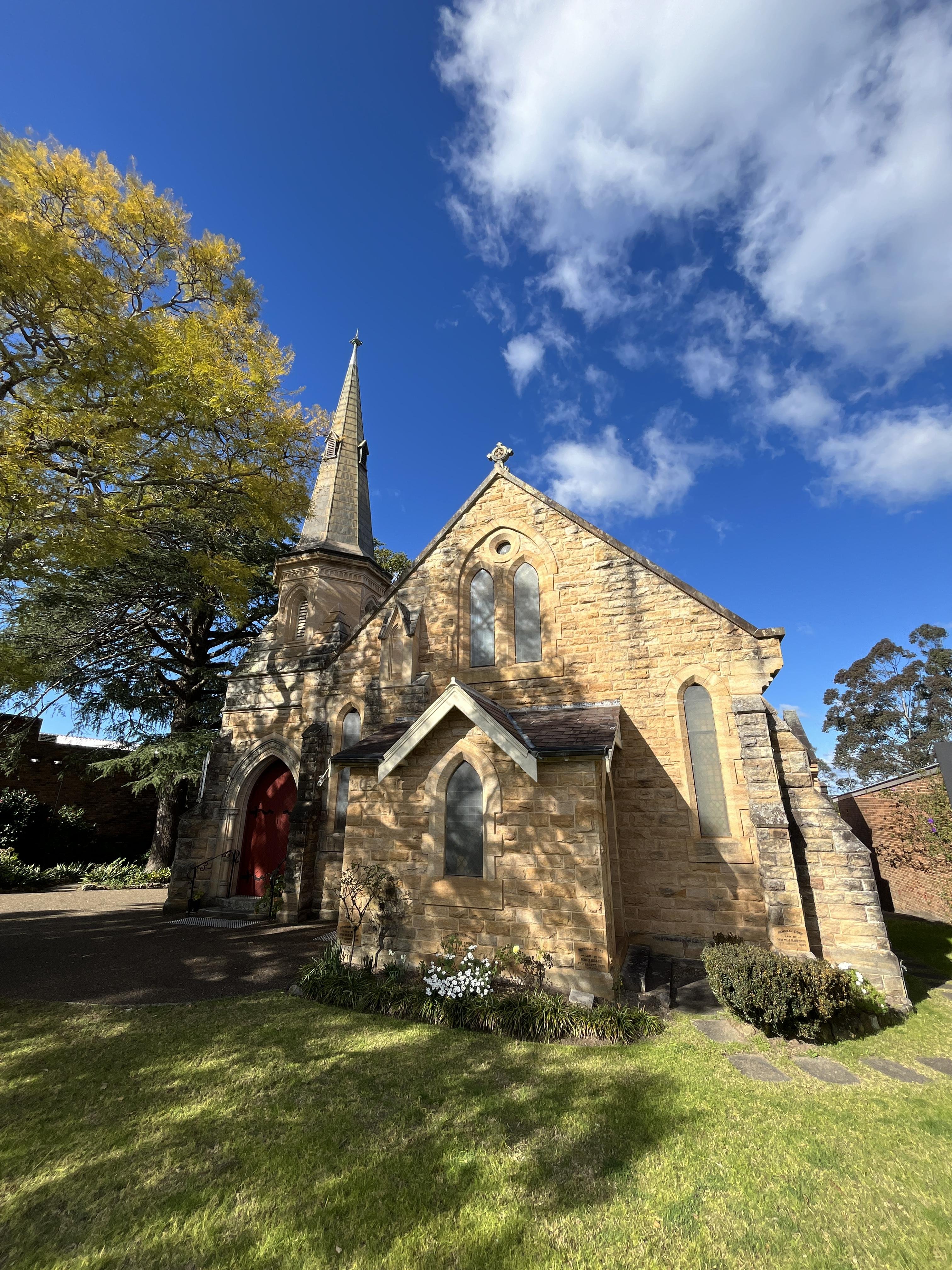 Stone church with tall, pointed steeple and red door, set against a bright blue sky with fluffy clouds. Yellowing tree on the left, casting shadows on the building. Foreground includes well-maintained grassy area and white flowers.