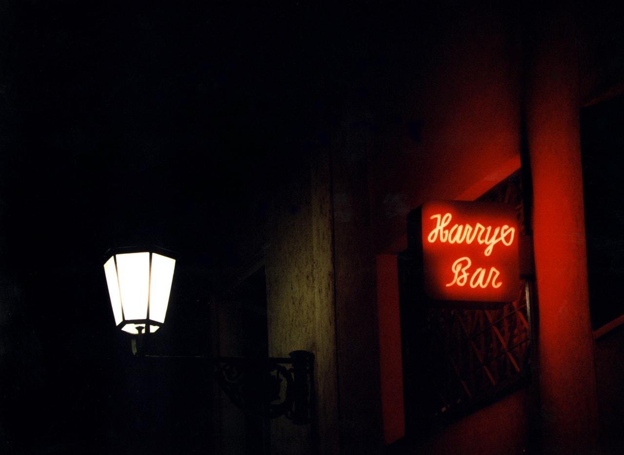 Dimly lit street with a white lantern on the left and a red neon sign on the right reading "Harry's Bar." The scene is predominantly dark, creating a contrast with the illuminated elements.