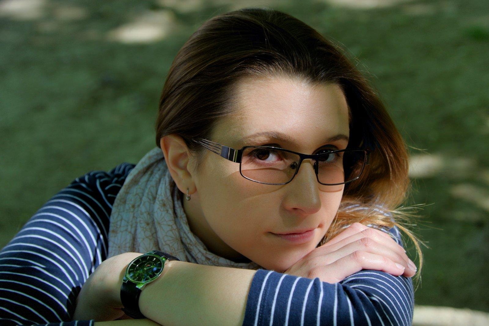 Gros plan d’une jeune femme brune portant un haut à manches longues bleu marine avec des trait horizontaux blanc, des lunettes, les bras croisés posés sur un support hors cadre et regardant la caméra d’un air très doux