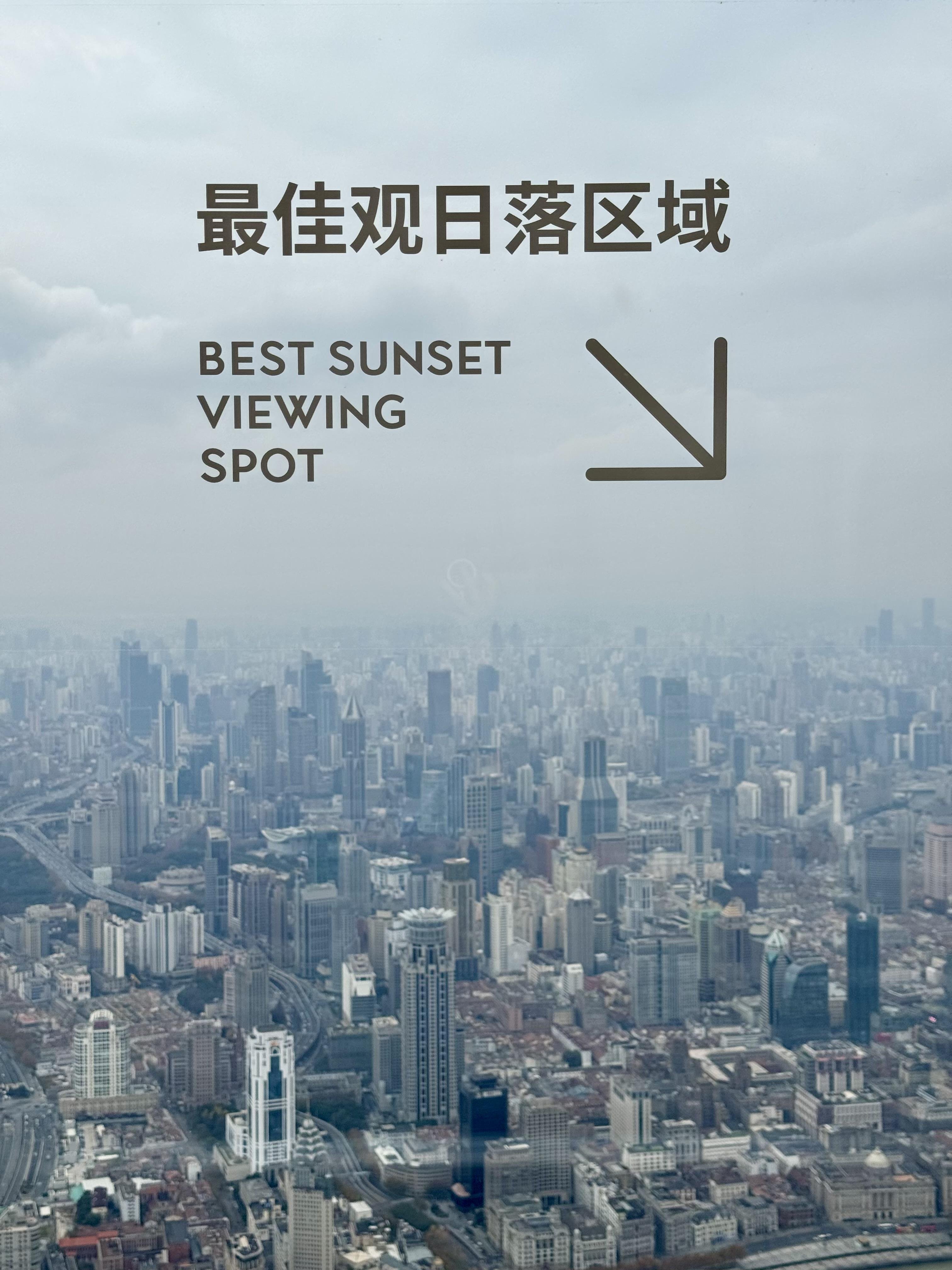 The image shows a cityscape with numerous tall buildings under a cloudy sky. Text in both Chinese ("最佳观日落区域") and English ("BEST SUNSET VIEWING SPOT") is overlayed, with an arrow pointing towards the right, indicating a good location for watching the sunset.