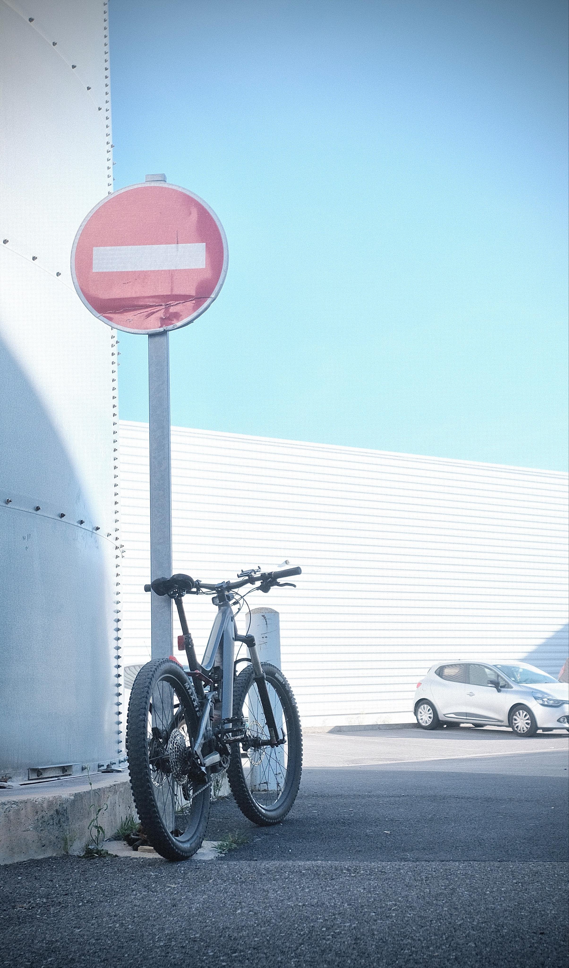 An industrial area with blue sky no clouds and harsh bright sunlight. In the picture’s foreground is shadow in which an electric fully is chained to a street sign „No entry for
vehicular traffic“. We are watching the scene from behind the bike looking at the car in the background. In the sun light background is a car with combustion engine parked.