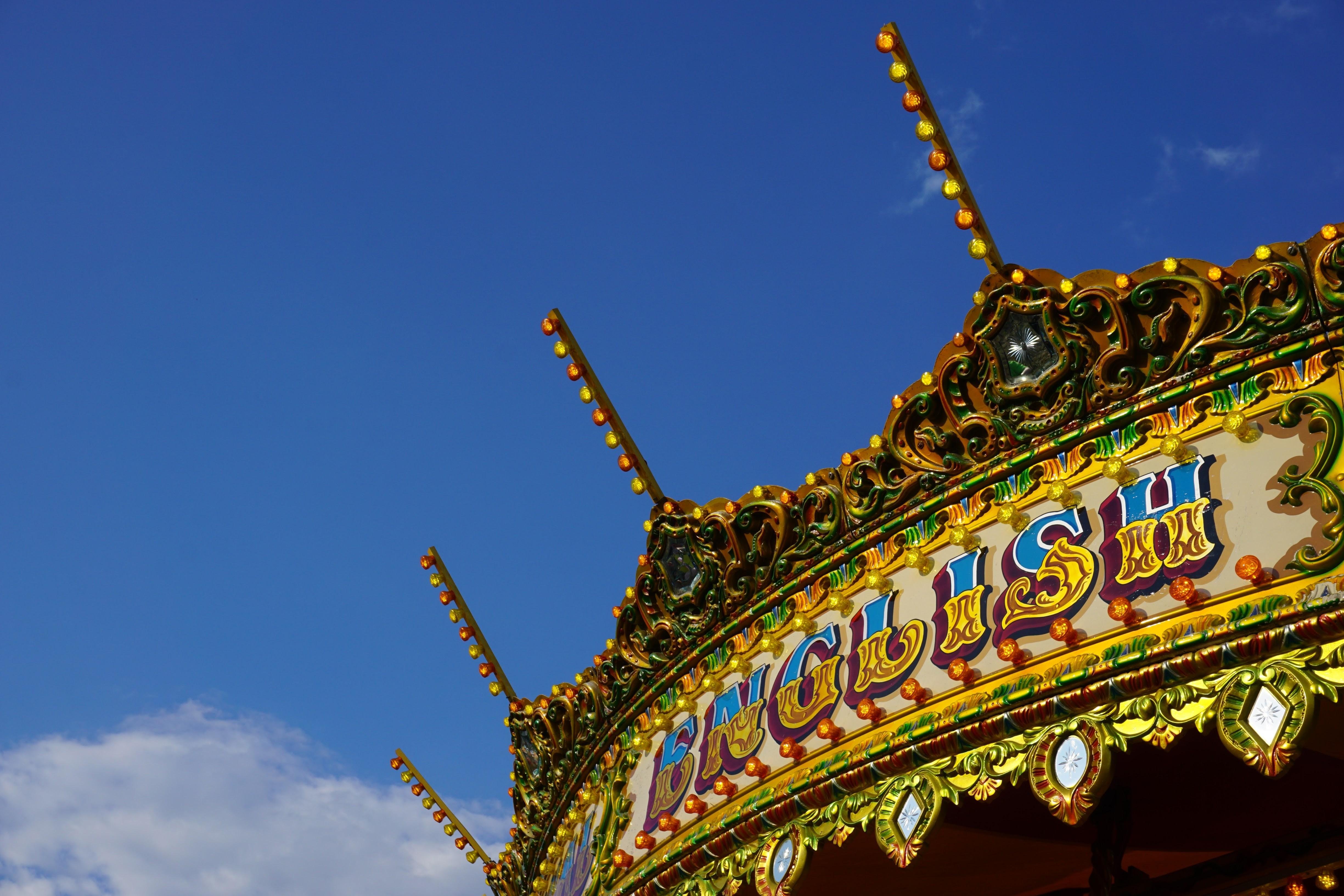 The image shows the ornate top of a traditional carousel, featuring intricate decorations and lights. The word "ENGLISH" is prominently visible in decorative, colorful letters. The sky is bright blue with a few clouds.