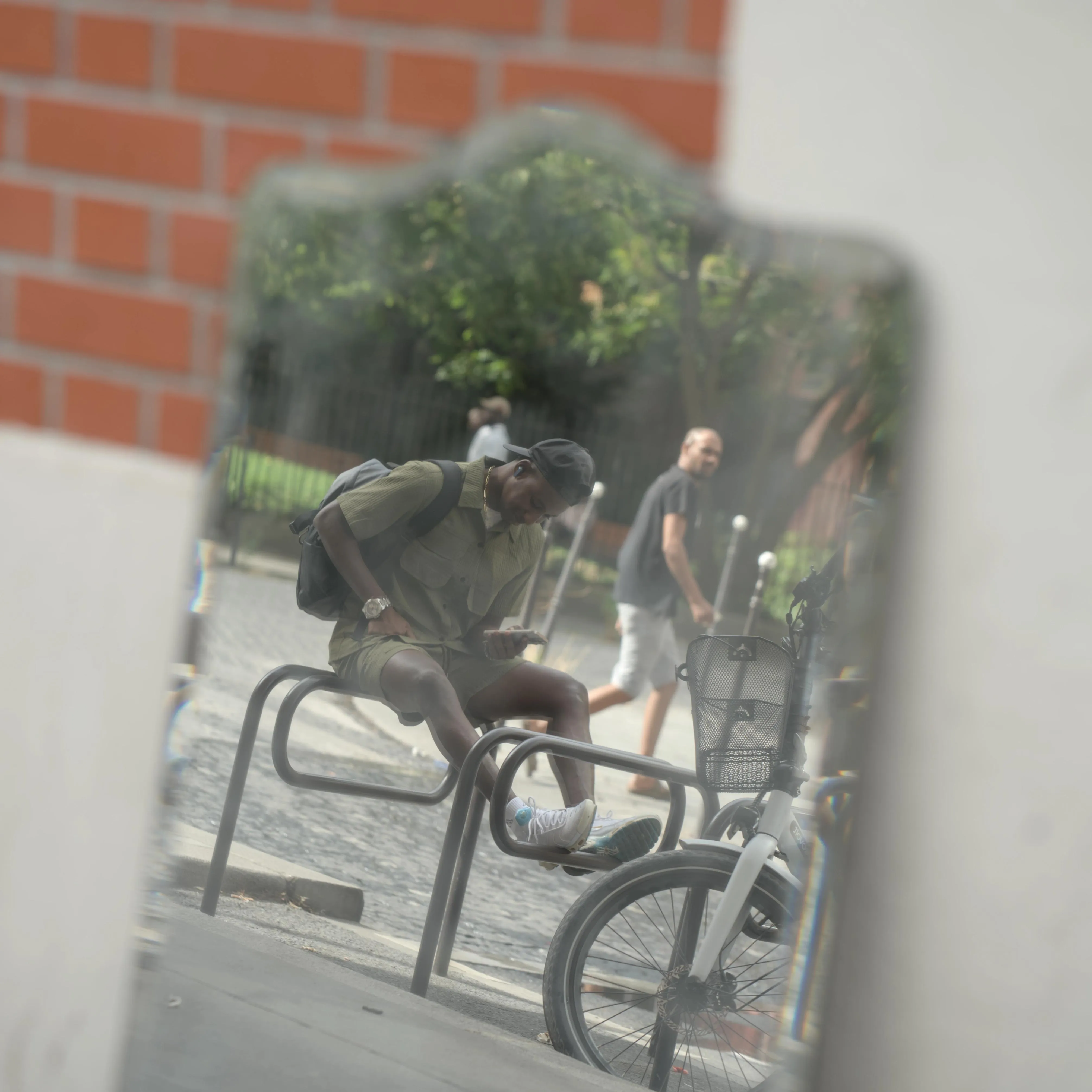 Photograph of a mirror on the wall of a streetside building showing the reflection of a man sitting on top of a bike rack on the sidewalk, with his feet resting on the next bike rack. The man is wearing a black baseball cap, a black backpack, black earbuds and an assorted green shirt and shorts. He is concentrated on his smartphone, slouching over it. Behind him, another man is walking on a pedestrian crosswalk and turning his head in the direction of the sitting man, mirror and camera. In the background of the reflection, there is greenery from a park on the other side of the street. The outer edges of the mirror and the brick wall the mirror is leaning on are blurry from the depth of field, while the reflection of the man sitting on the bike rack is in focus. The edges of the mirror have chromatic aberrations.
