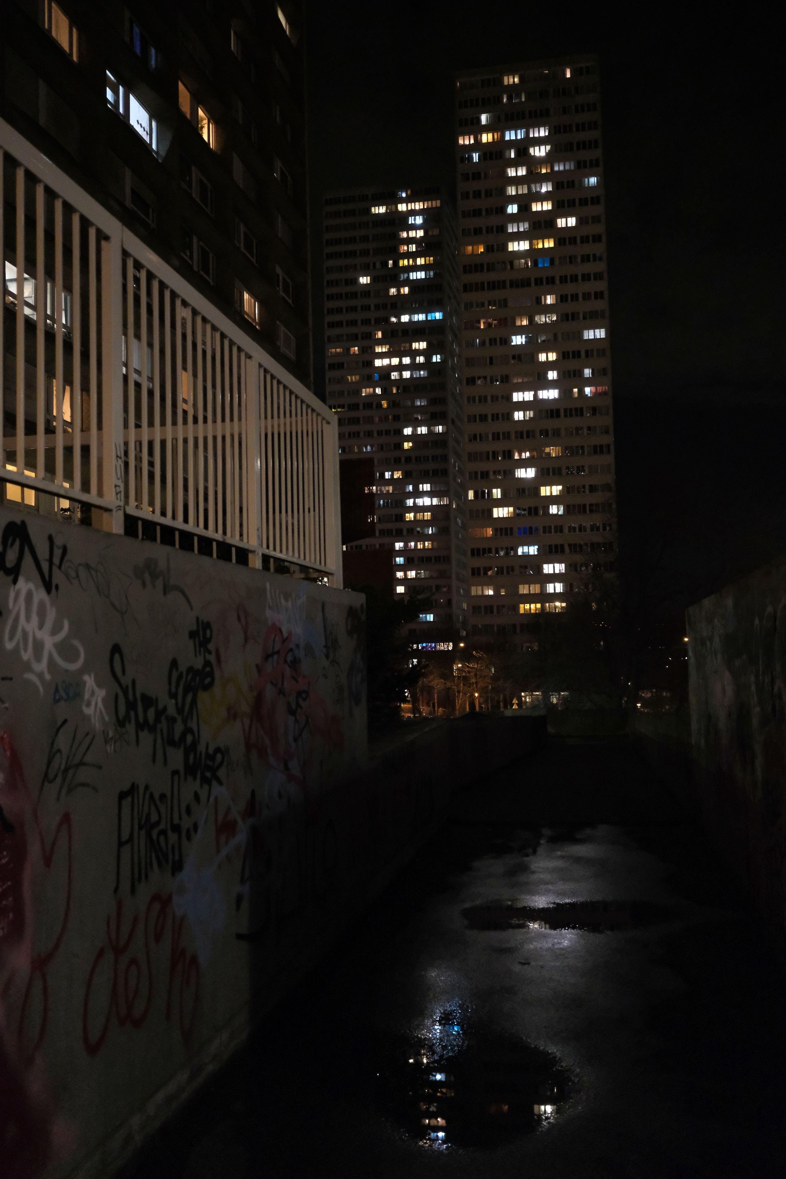 Photo format portrait, de nuit, d’une allée piétonne sombre et étroite, le mur de gauche plein de graffitis, une barrière au sommet, le sol humide avec des flaques d’eau, des hautes tours d’immeubles avec leur lumières inégales aux fenêtres