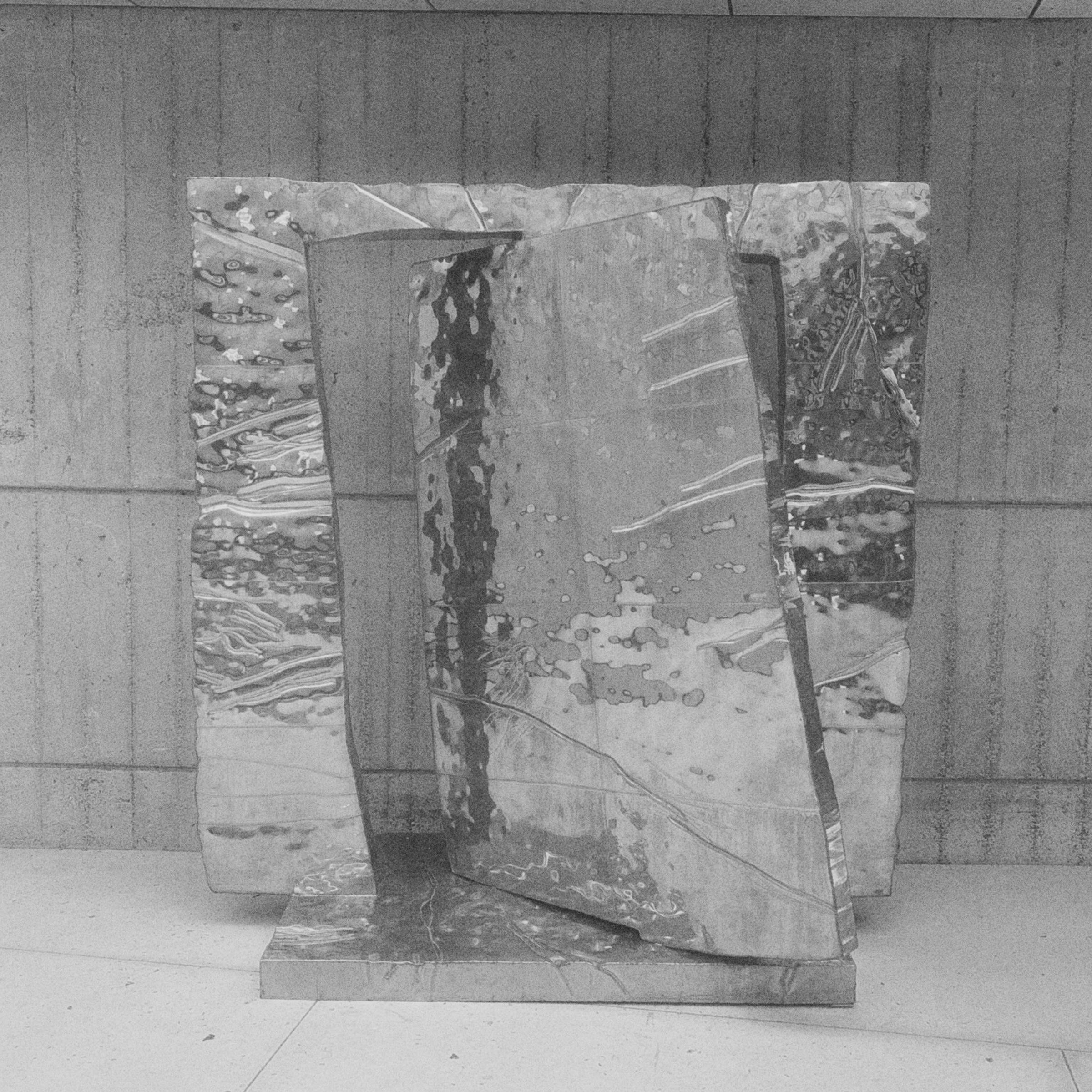 Three tall, irregular metal panels with reflective, rippled surfaces stand on a rectangular base in front of a concrete wall, with the center panel angled forward and seams visible between the panels.