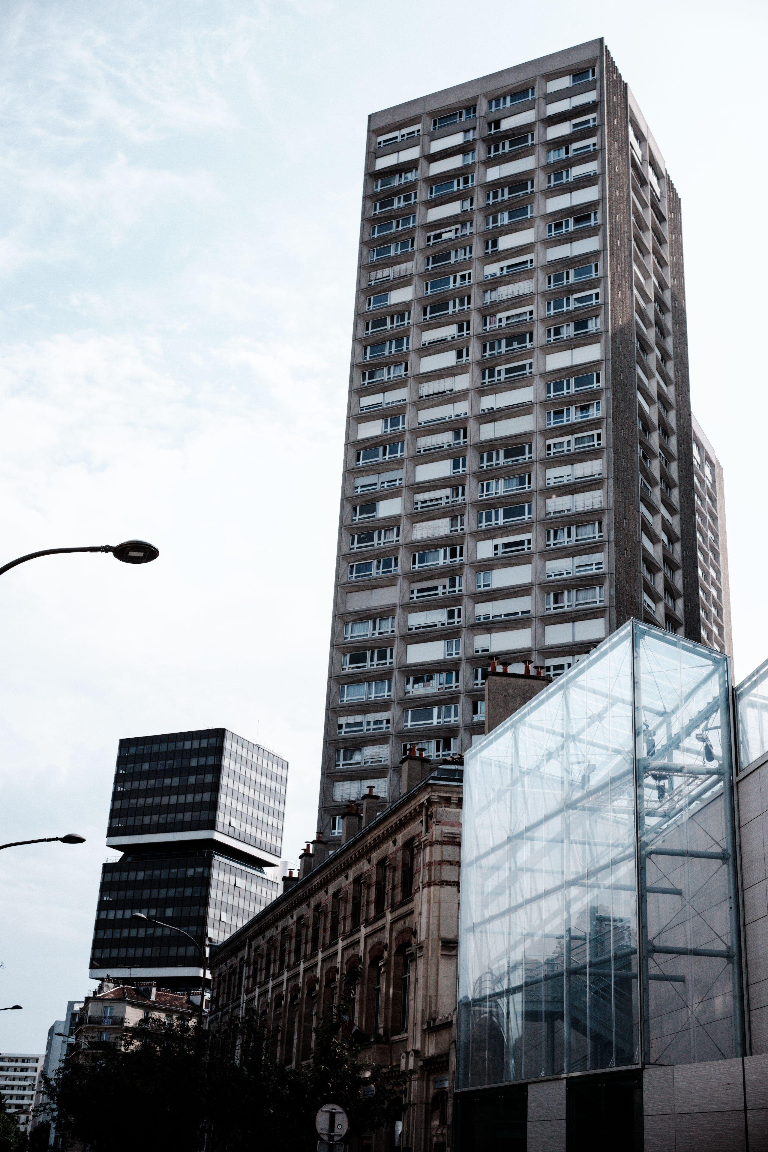 Photo verticale en noir et blanc. Au premier plan, un petit bâtiment en verre, suivi d’un autre plus classique. Derrière eux, une tour d’habitation de plusieurs dizaines d’étages, et la tour de la fac de Tolbiac