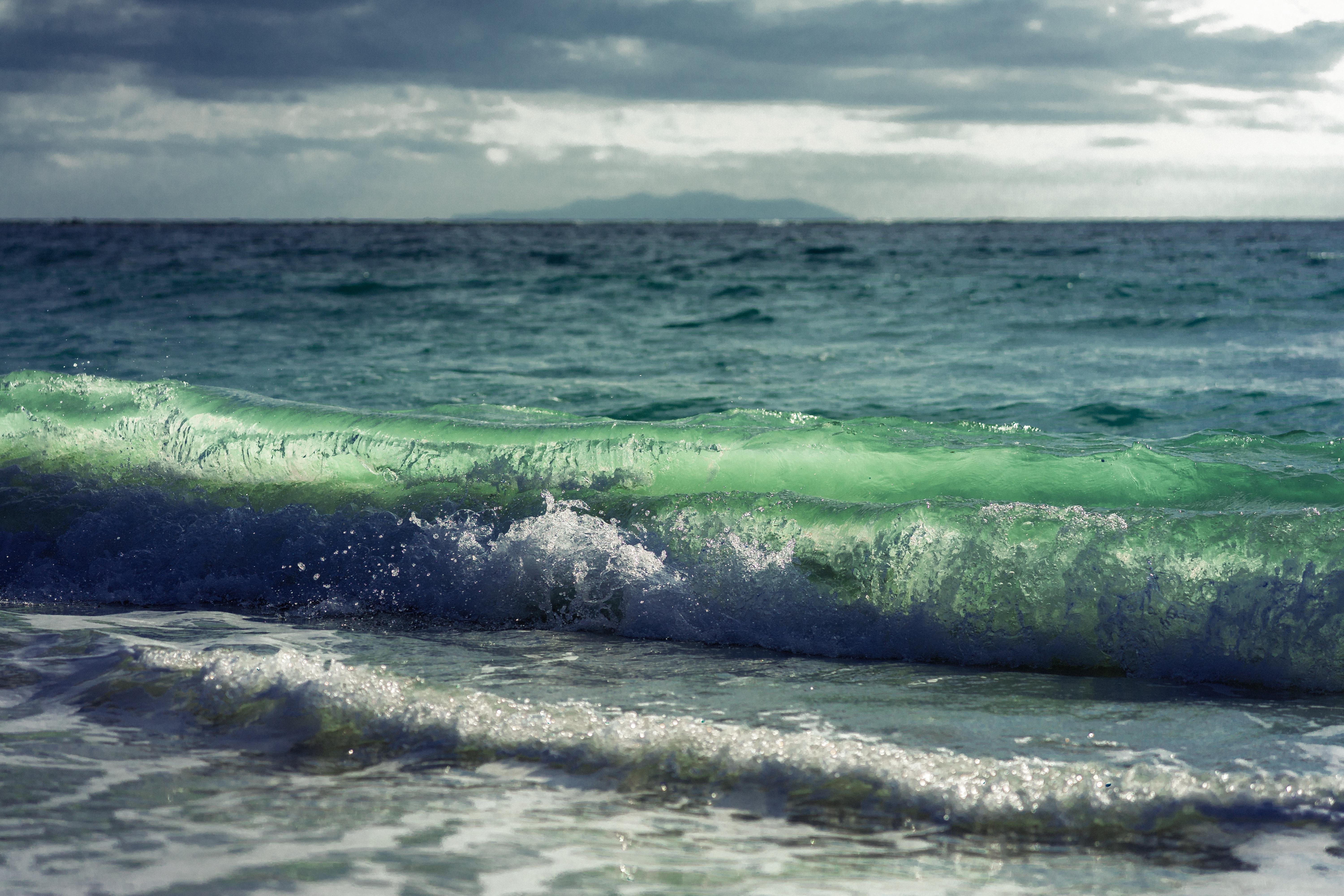 The image shows a view of the ocean with a wave crashing in the foreground. The water is a vibrant greenish-blue, and the sky is cloudy, with a distant landmass visible on the horizon. The scene captures the dynamic motion of the wave and the expansive ocean.