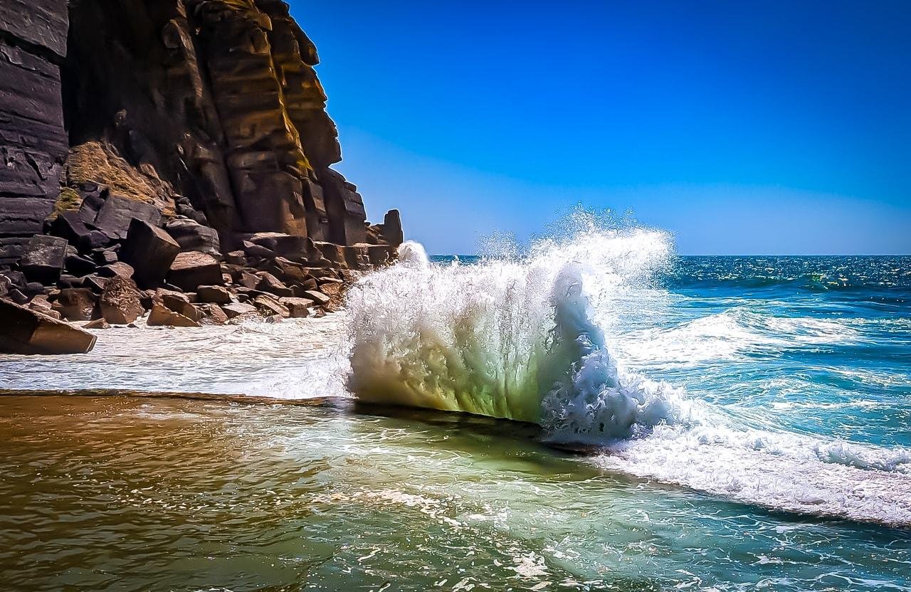 Large wave crashing against a cliffside on a sunny day, with clear blue skies and turquoise ocean water. Rugged rocks are visible along the shoreline, and foam from the wave spreads across the sand.