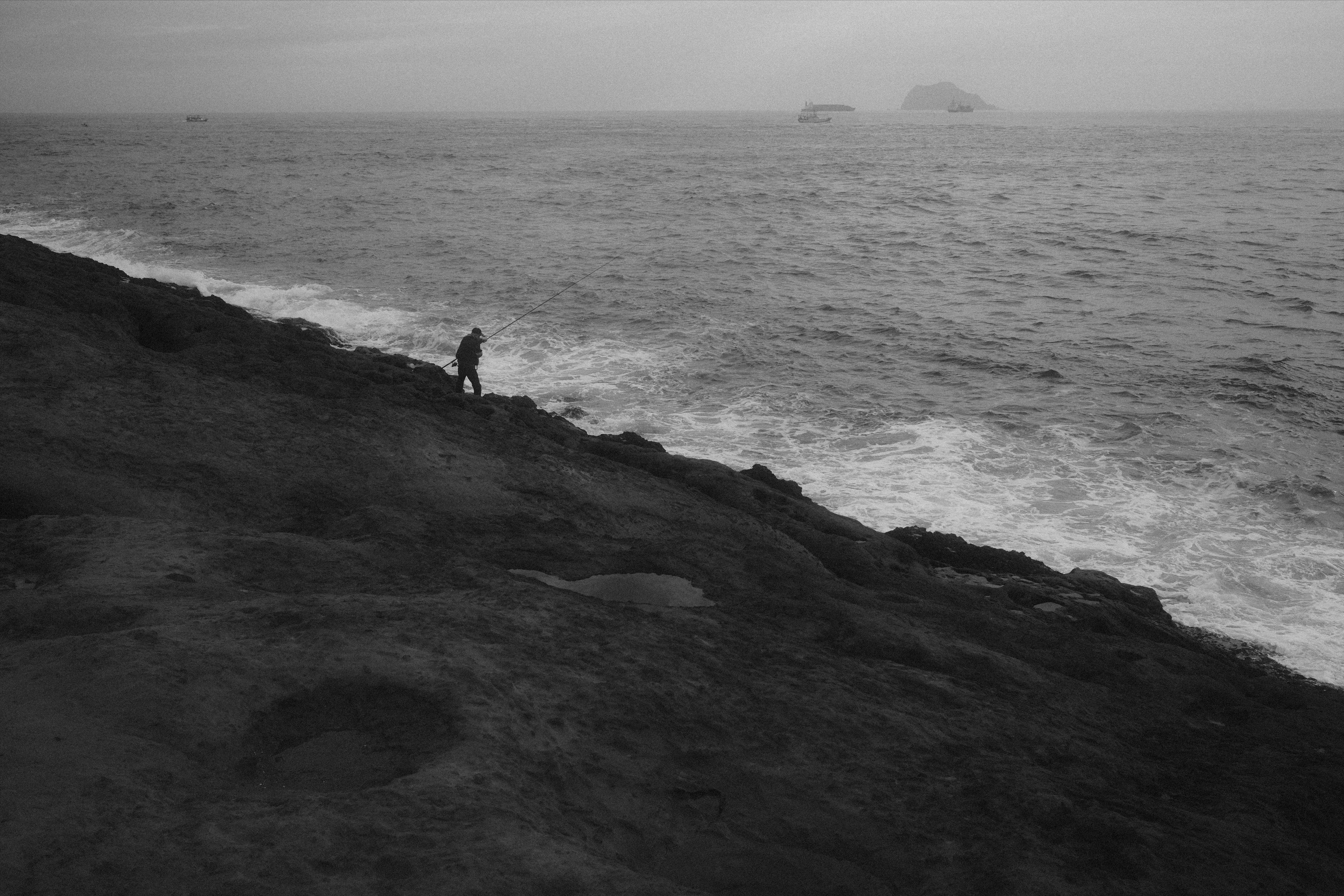 Person fishing from a rocky shoreline in the foreground, with waves breaking along the coast and an overcast sea extending to the horizon with several small boats and a distant island silhouette.