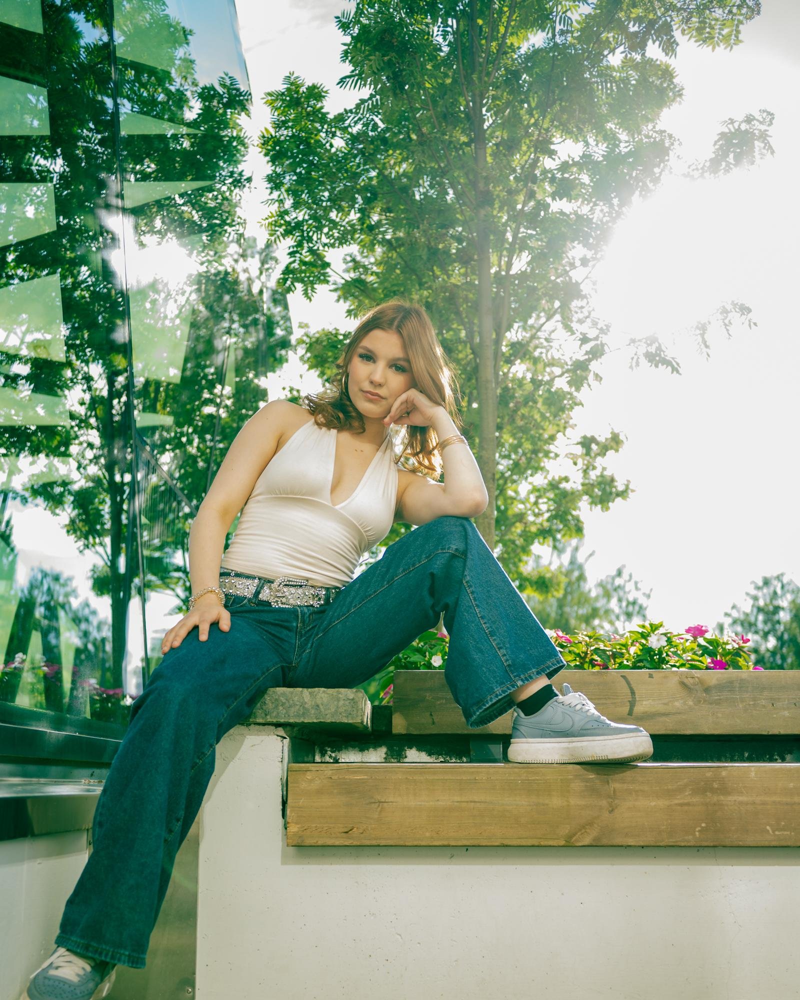 Young woman with shoulder-length reddish-brown hair sits casually outdoors on a raised wooden ledge. She is wearing a white sleeveless top, blue jeans, and light gray sneakers, with a reflective building surface on the left and a tall tree with greenery in the background. Bright sunshine filters through the foliage.