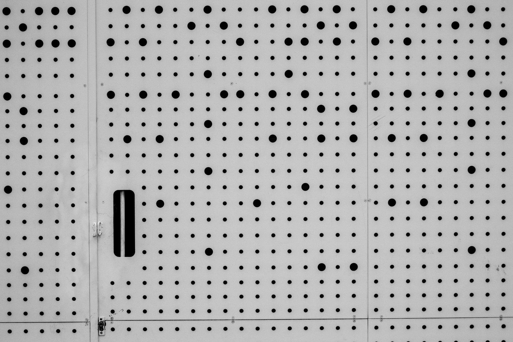 Photo horizontale en noir et blanc. Une surface blanche pleine de trous de 2 tailles différentes. Une poignée est présente nous faisant comprendre que c’est une porte