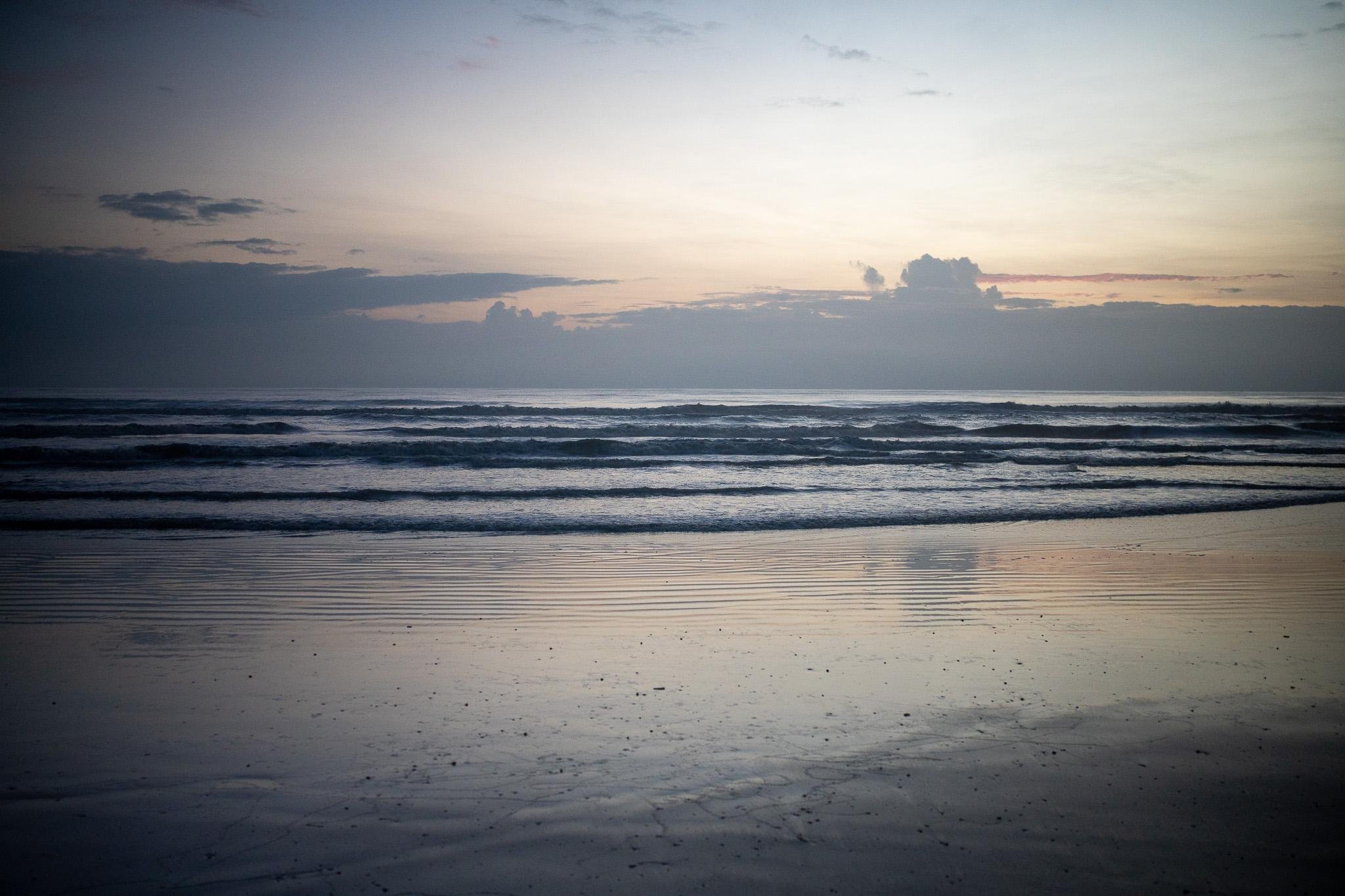 The image shows a beach at either sunrise or sunset. The sky has soft colors, and there are clouds on the horizon. The water is calm with gentle waves, and the sand in the foreground reflects the light from the sky.