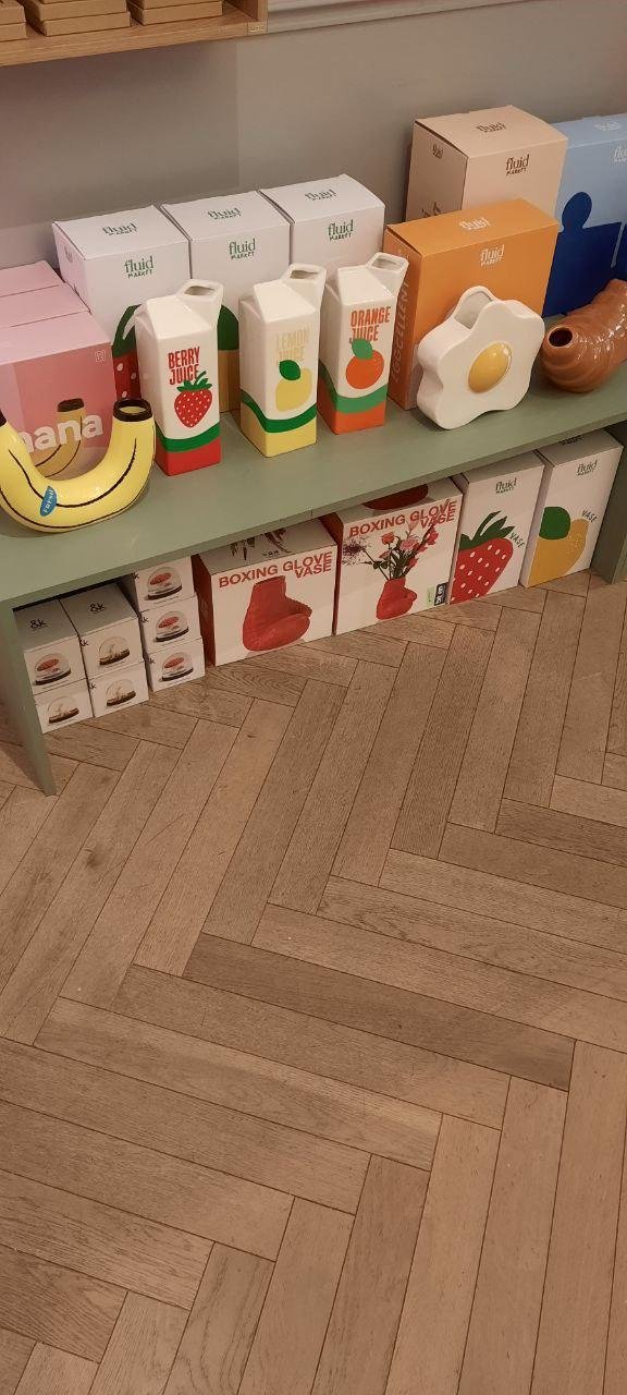 Die Bilder zeigen Regale mit verschiedenen Objekten die wie alltägliche dinge aussehen.
- Vasen die wie Tetrapackungen aussehen mit den Aufschriften wie "Berry Juice," "Lemon Juice," und "Orange Juice."
- Vasen die aussehen wie Lebensmittel: Corossaint, Banane, Spiegelei, Ananas, Chinakohl
- Vase als Boxhandschuh
Decken, Kissen, Taschen hängen im Schaufenster sowie schlichte und minimalistische Kukuksuhren.