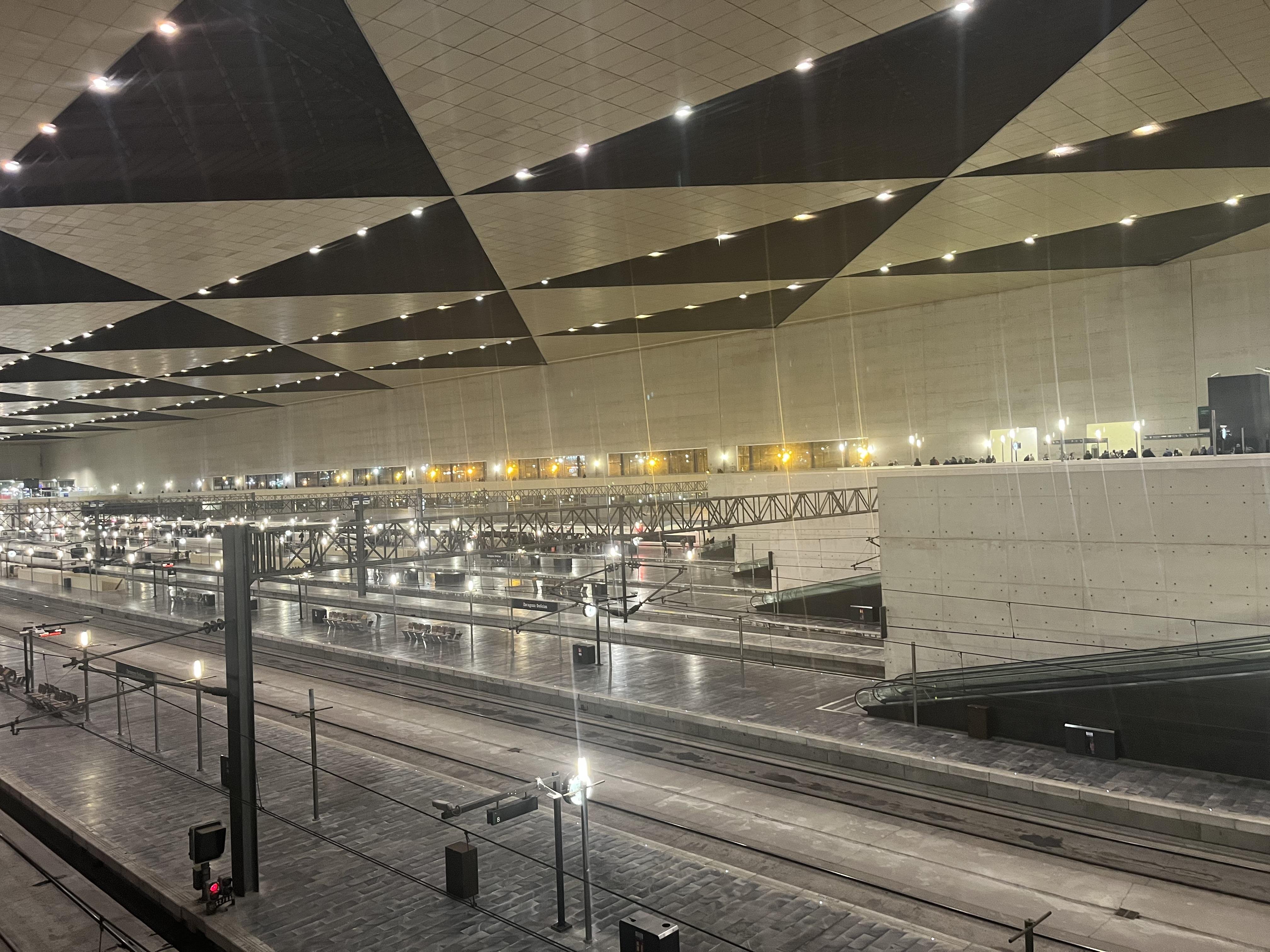 This image shows an inside view of a large, modern train station. There are multiple train tracks and platforms visible, with an expansive and well-lit ceiling featuring geometric patterns. The station appears to be spacious and designed with a minimalist architectural style.