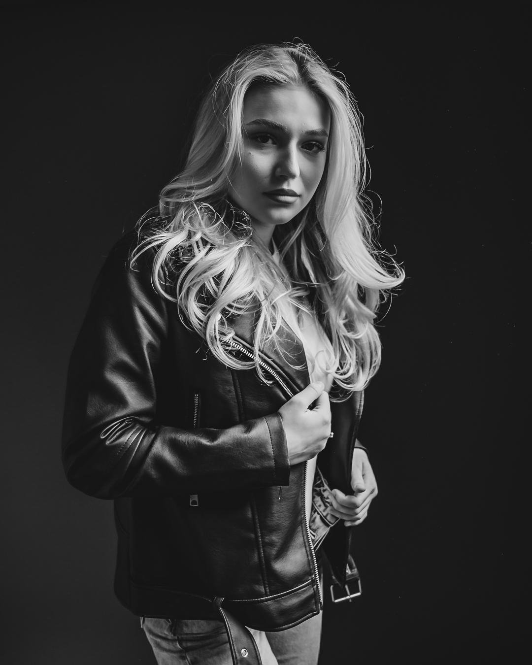 The image depicts a black-and-white portrait of a person with long, wavy hair wearing a leather jacket. The person is posing against a dark background.