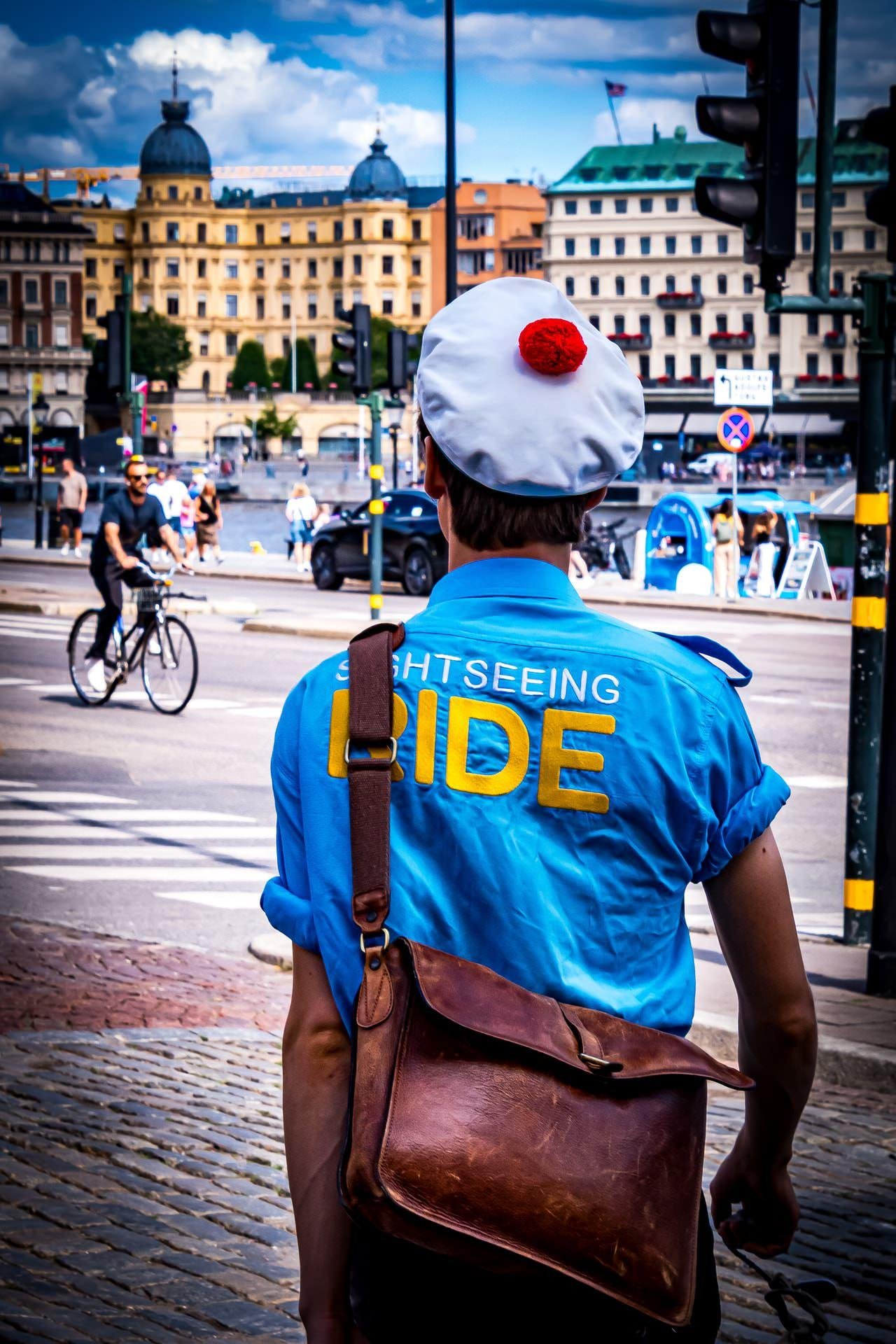 Person wearing a blue shirt with "Sightseeing Ride" printed on the back, and a white hat with a red pompom. A brown leather bag is slung over their shoulder. They stand at an intersection with cobblestone pavement. In the background, people walk and cycle near a street lined with historic buildings under a partly cloudy sky.