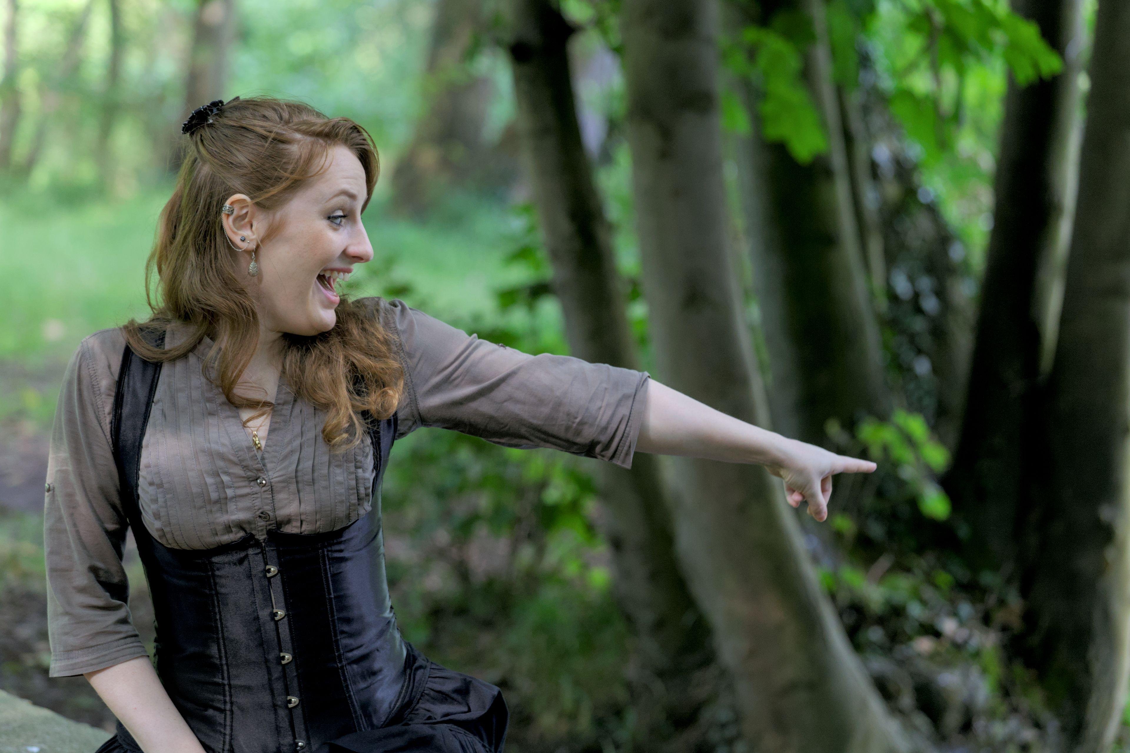 Photo horizontale, montrant une jeune femme rousse au cheveux long élégamment coiffé, portant un chemisier marron et par dessus, un corset noir. Elle pointe du doigt quelque chose hors cadre avec un grand sourire de surprise