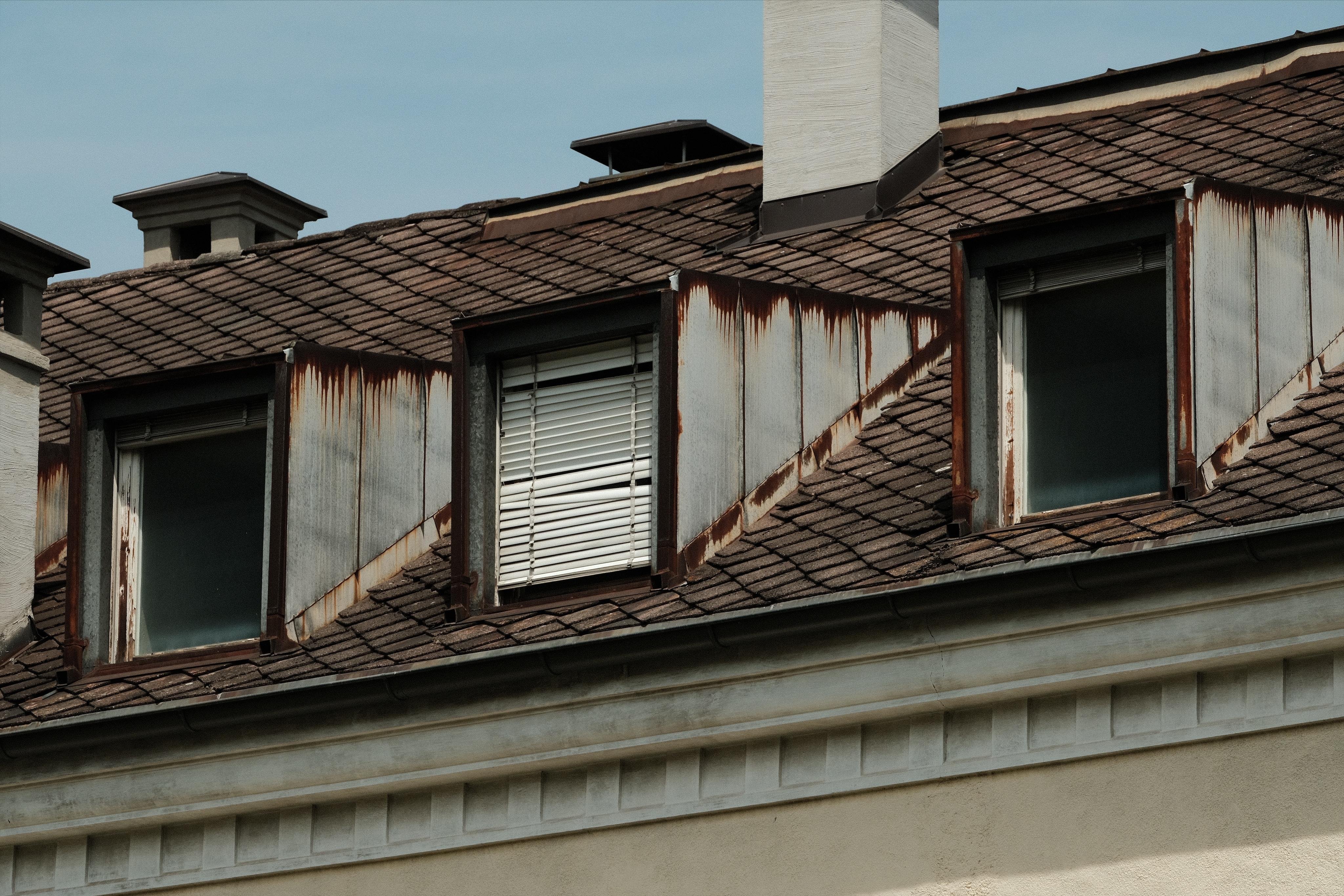 Sloped roof with three dormer windows, rust-streaked metal siding around the dormers, a partially closed and broken shutter in the center window, and a white chimney against a clear sky.