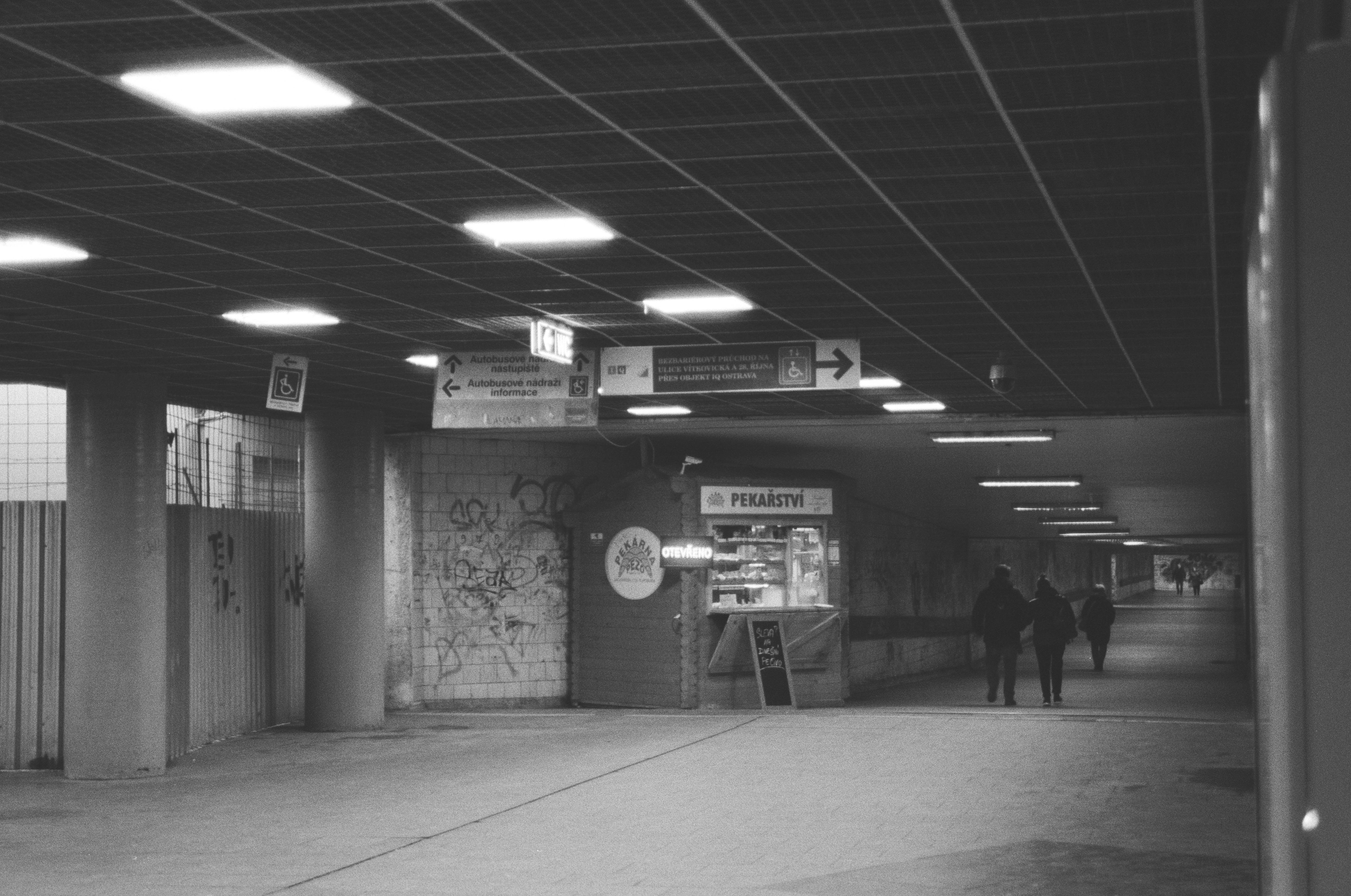 A monochrome photo from within a pedestrian underpass with long tunnel ahead, a small food stand on the left side and some ongoing construction on the far left. There is a row of square light fixtures on the ceiling and some people in the distance