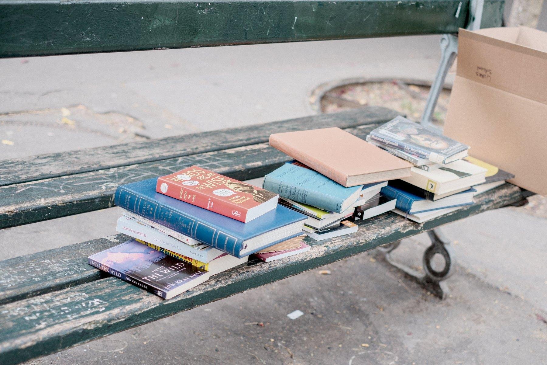 Photo horizontale. Sur un banc parisien vert, des livres posés dessus en petit tas, et on voit un bout de carton posé sur le banc.