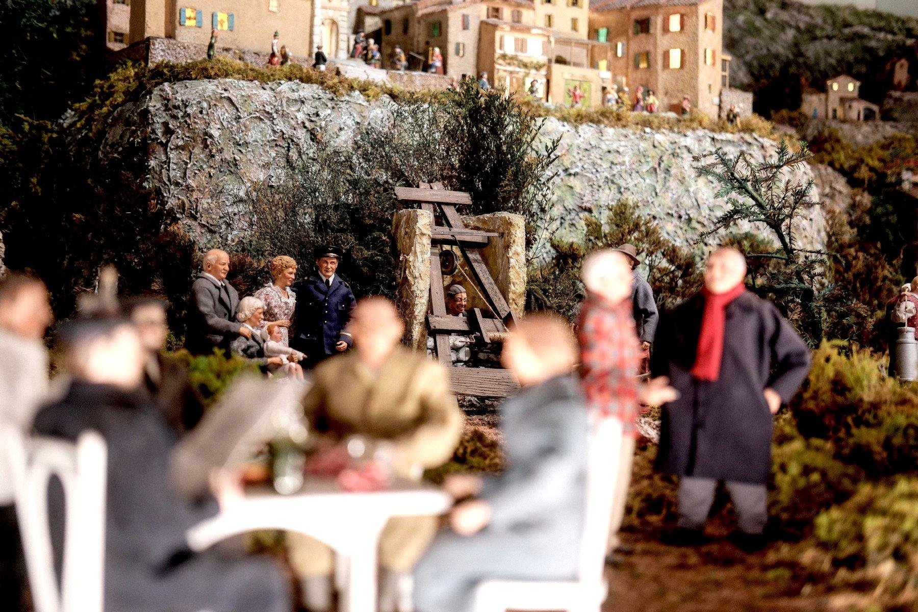 Photo horizontale. Photo rapprochée sur un diorama, premier plan flou avec des personnages assis à une table à gauche, puis toujours flou, 2 personnes debout sur la droite, et net, un personnage dont la tête dépasse de l’entrée d’un puit de mine