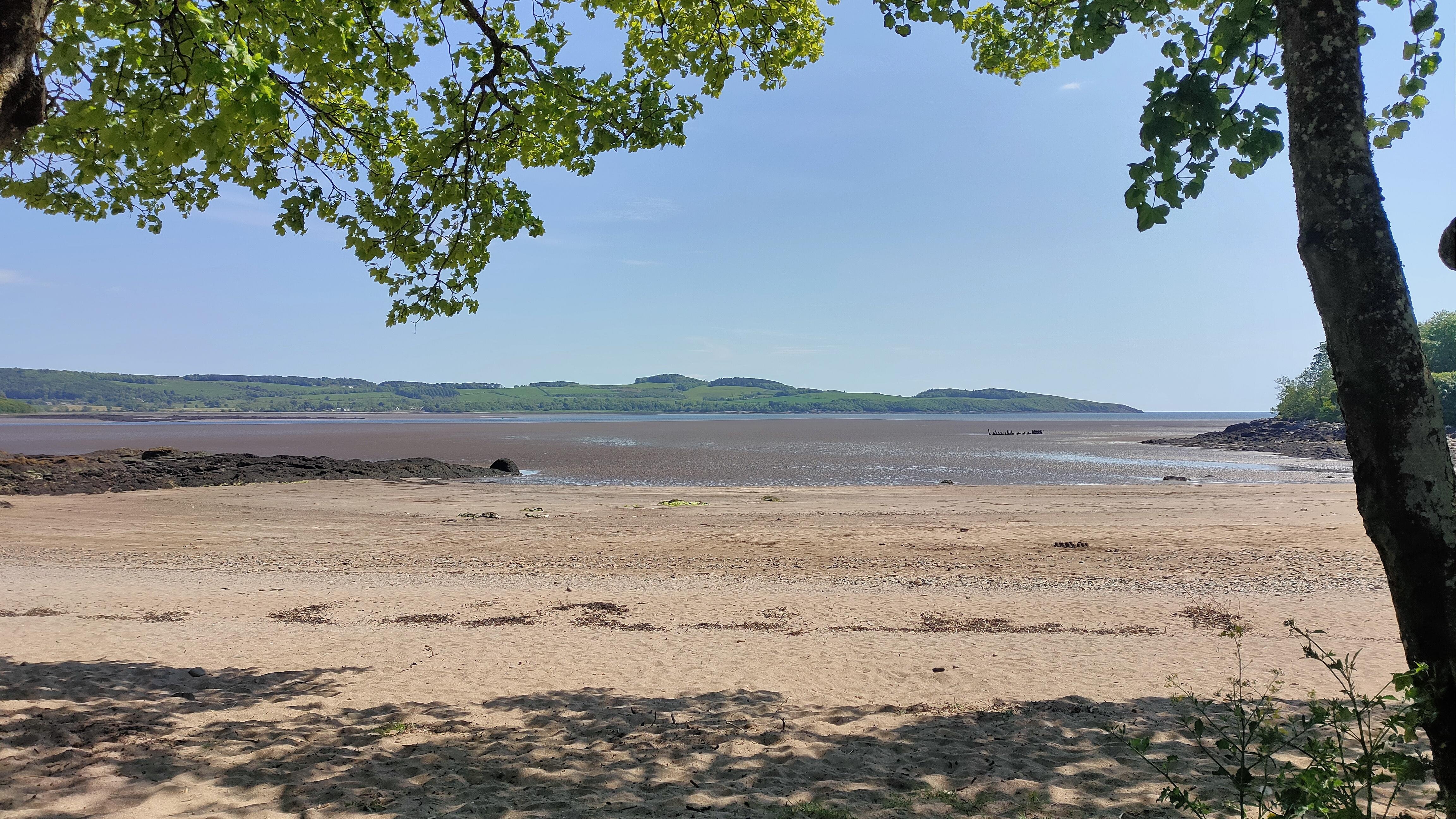 The image shows a serene beach scene with sandy shores in the foreground and calm water leading to a distant, hilly landscape. There are trees framing the top of the picture, providing shade and a natural border. The scene appears peaceful and inviting, likely on a clear day.