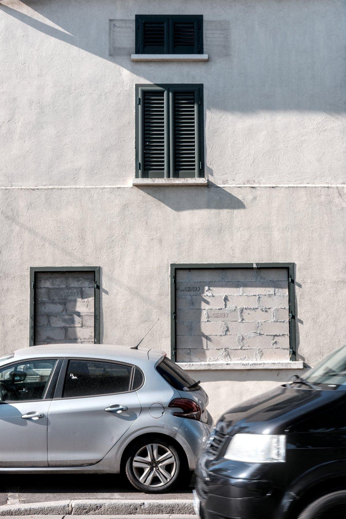 Photo verticale. Façade d’une maison au mur blanc avec des cadres de fenêtres verts sombre. Tout en haut centré, une toute petite fenêtre avec juste en dessous une fenêtre normal, les deux ont leur volets verts fermés. Au rez-de-chaussé, 2 fenêtres de tailles différentes, murées. Une voiture grise garée devant, l’avant d’un van noir entre dans le cadre par la droite au premier plan