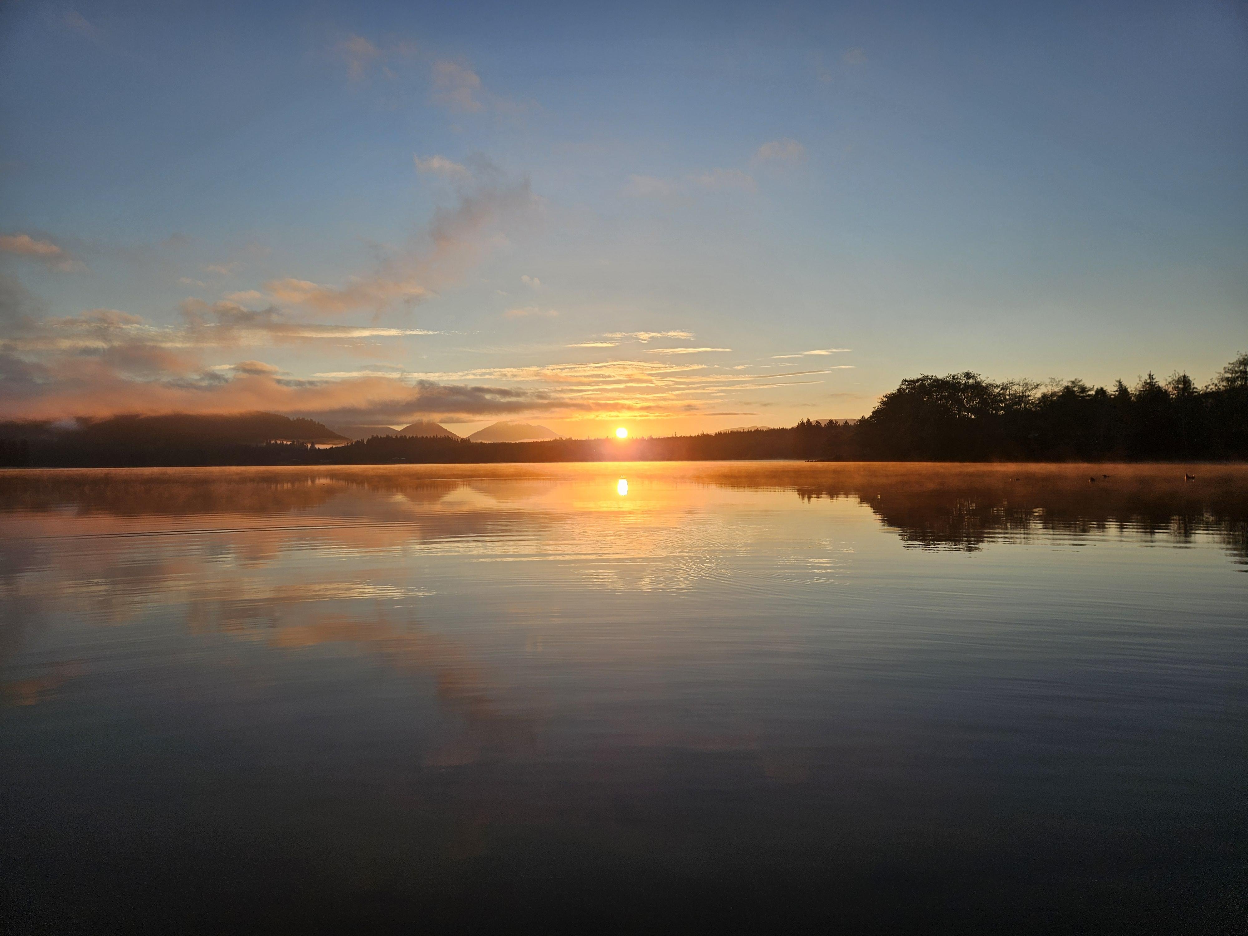 Low sun on the horizon over a calm lake, casting a bright reflection across the water; thin mist along the shoreline with silhouetted trees on the right and distant hills on the left under scattered clouds.