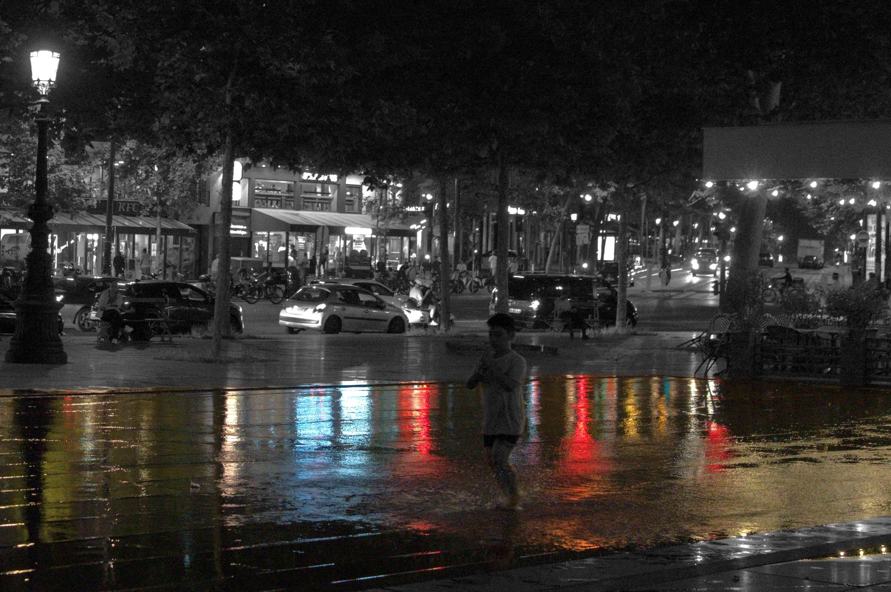 Photograph of a child running on a public square. He is facing 3/4 towards the camera. His hands are joined in front of him. Most of the surface of the square visible in the image is covered in water. The child's feet are making splashes and ripples in the water. City lights are reflected on the water surface. The surface of the water has a green coloration, reflecting the trees surrounding the square. The reflected city lights form bright red, yellow and blue columns on the water surface. The rest of the photograph is in monochrome, including the child.