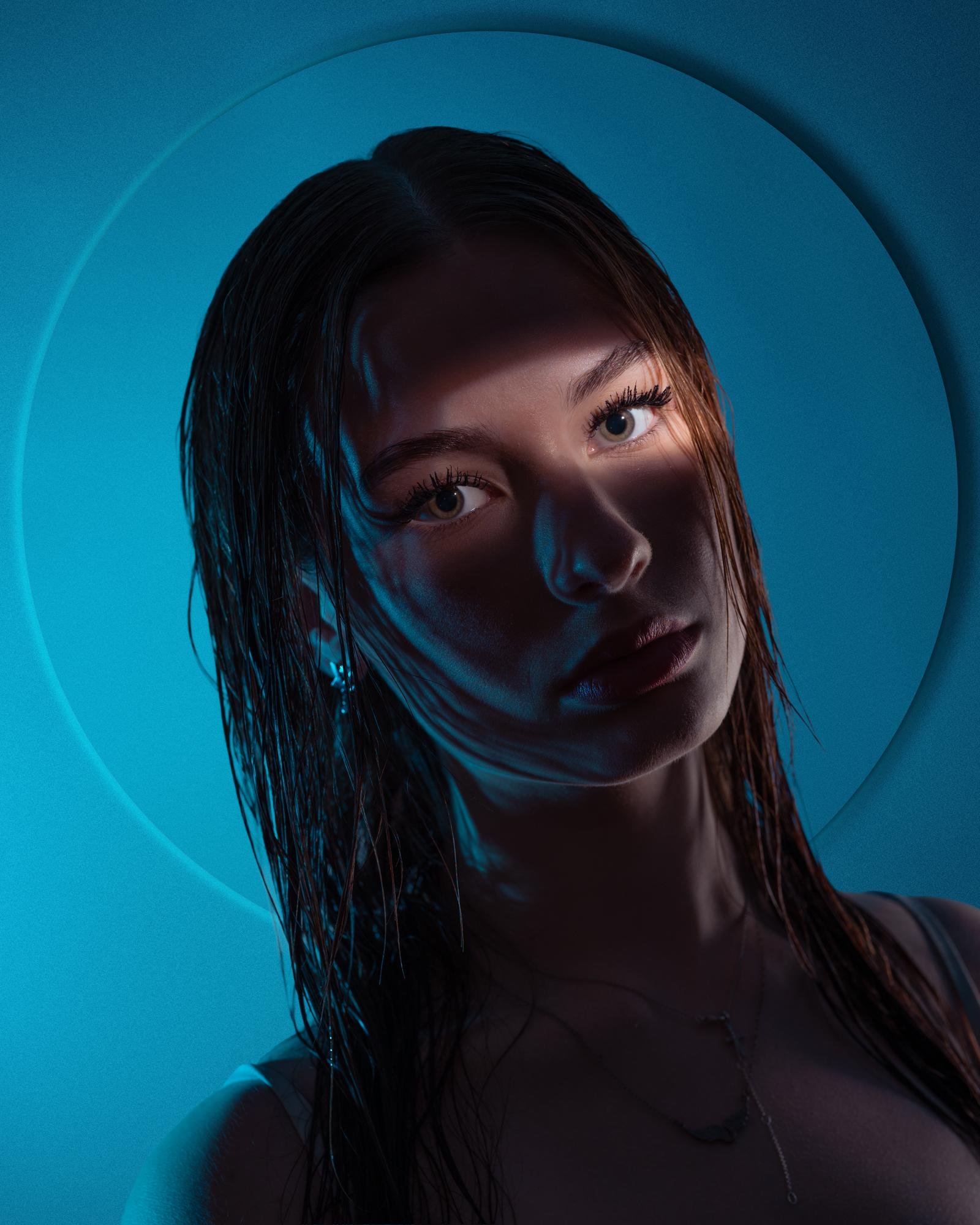 A woman with wet hair faces forward under dramatic blue lighting, creating shadows across her face. Her eyes are wide open, and she wears earrings and a necklace. A circular backdrop enhances the composition by casting a subtle halo effect around her head.