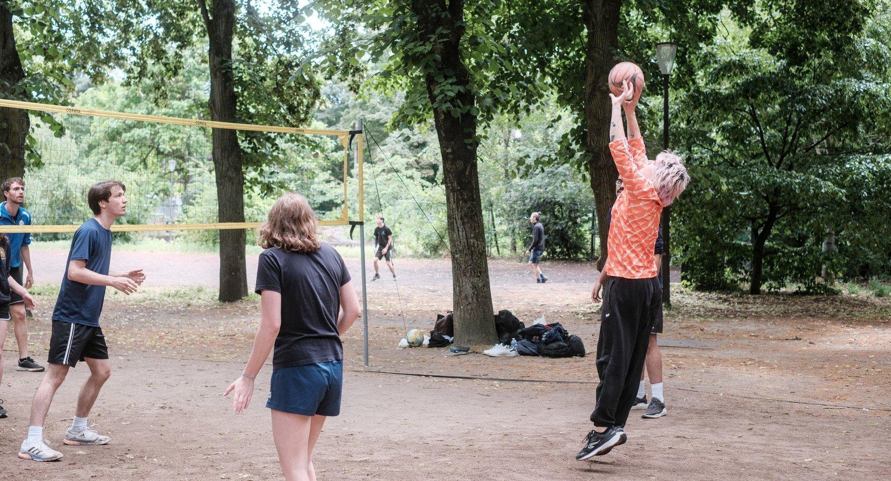 Photo horizontale. Une partie de volley dans un parc, sur la droite, une jeune femme aux cheveux violet, haut orange en plein saut pour renvoyer la balle du bout des doigts, sur la gauche, un jeune homme de son équipe en mouvement, au centre, une jeune femme de dos, en short, regarde l’action
